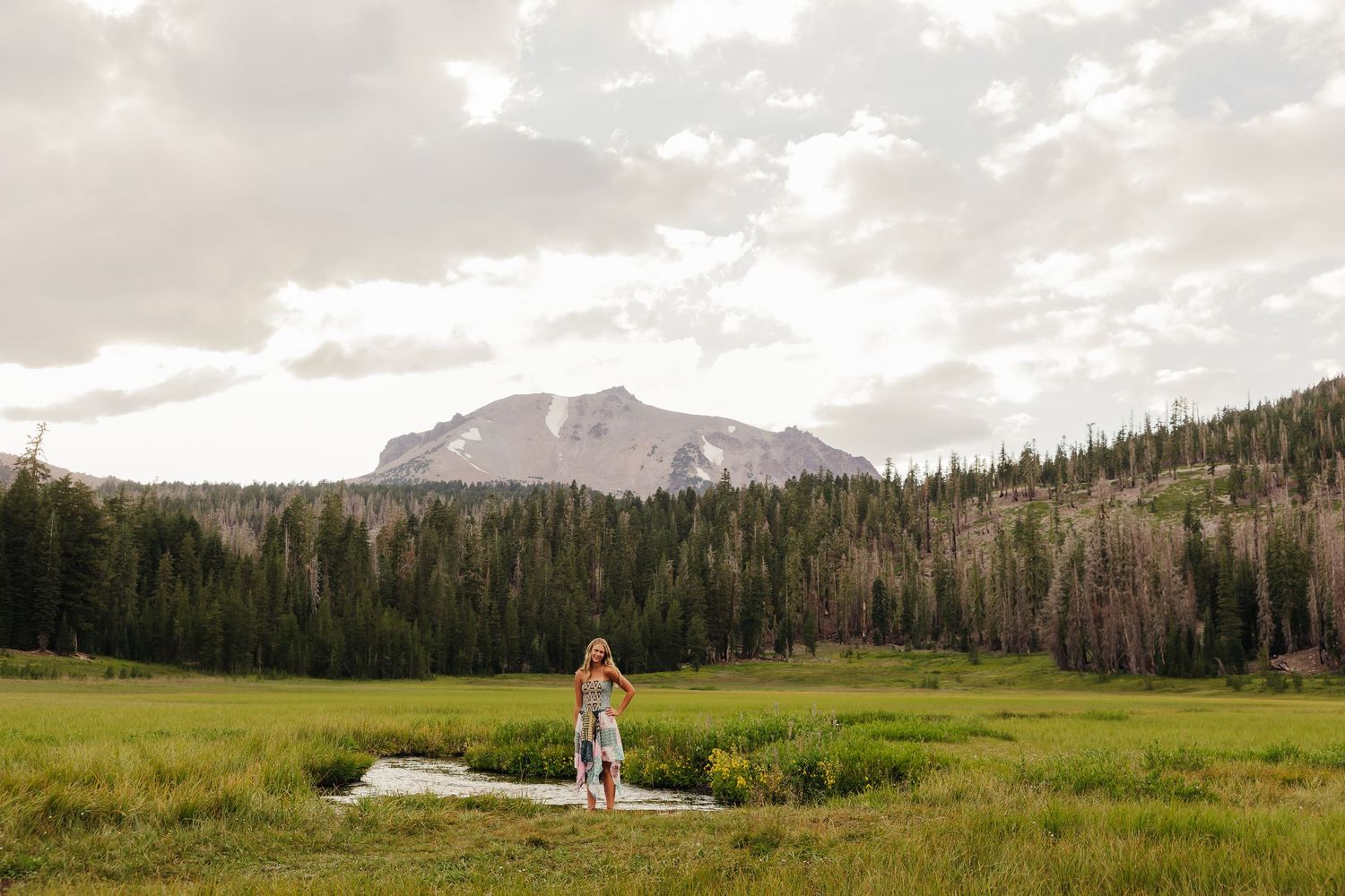 A couple stands in a lush green mountain meadow with a stream, surrounded by pine forests and a rocky peak.
