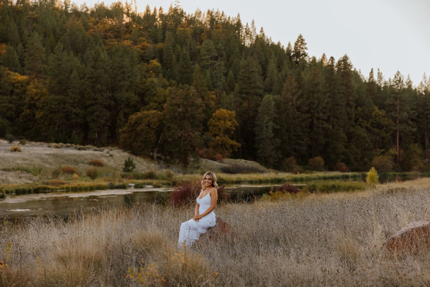 Woman in white dress poses in tall grass near lake with pine forest backdrop.