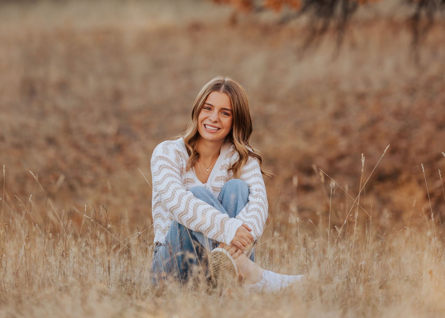 A woman sits in a golden field wearing casual denim and a light sweater.