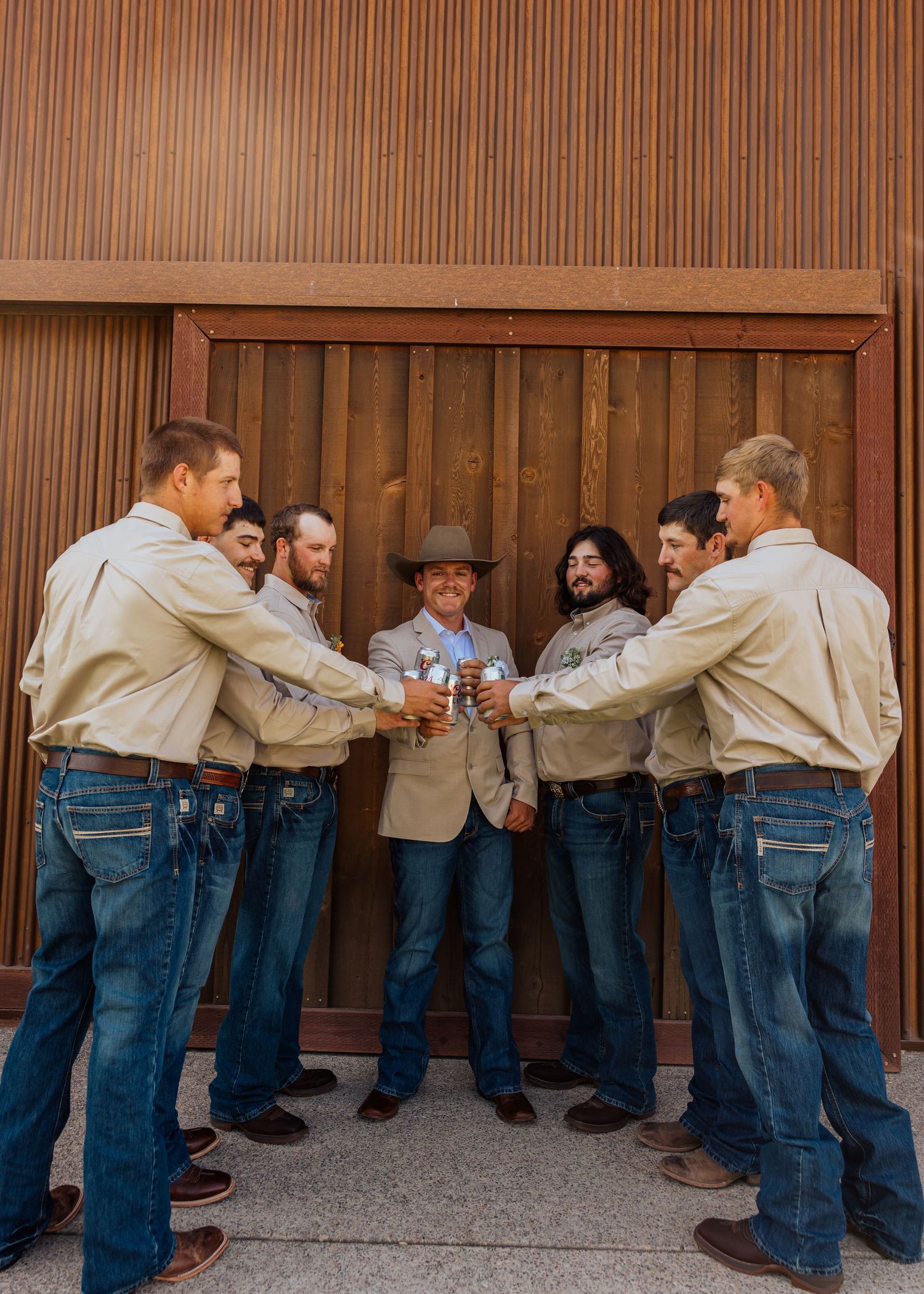 Groomsmen in matching tan jackets and jeans line up against wooden wall for creative photo.