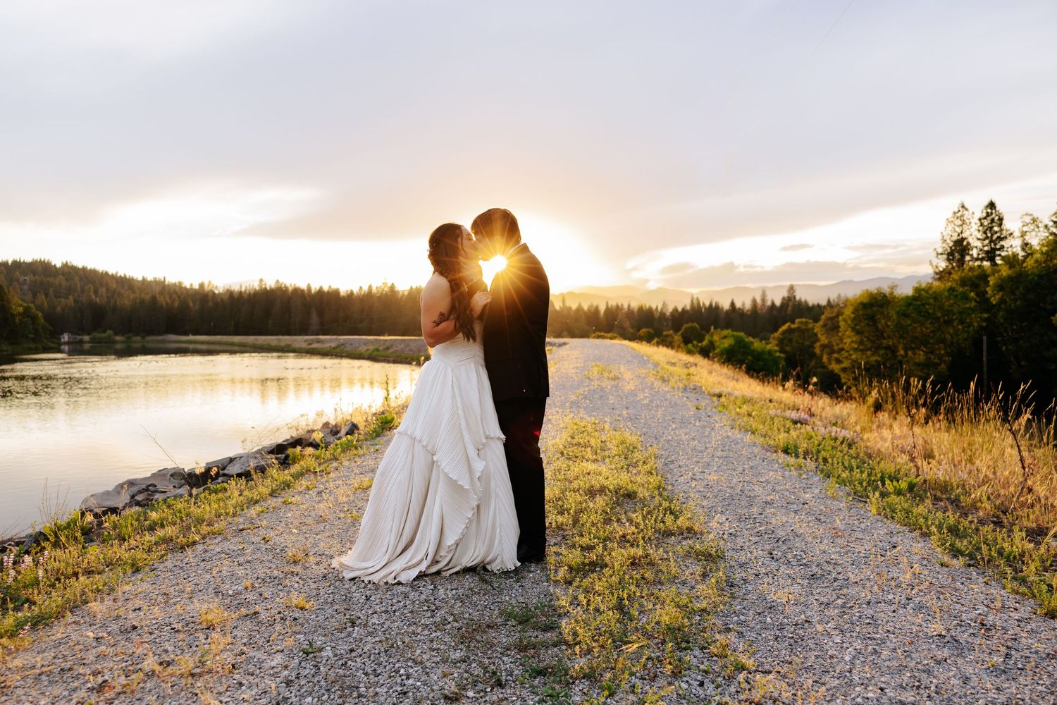 Bride and groom share a romantic sunset kiss on a gravel path beside a serene lake surrounded by trees.