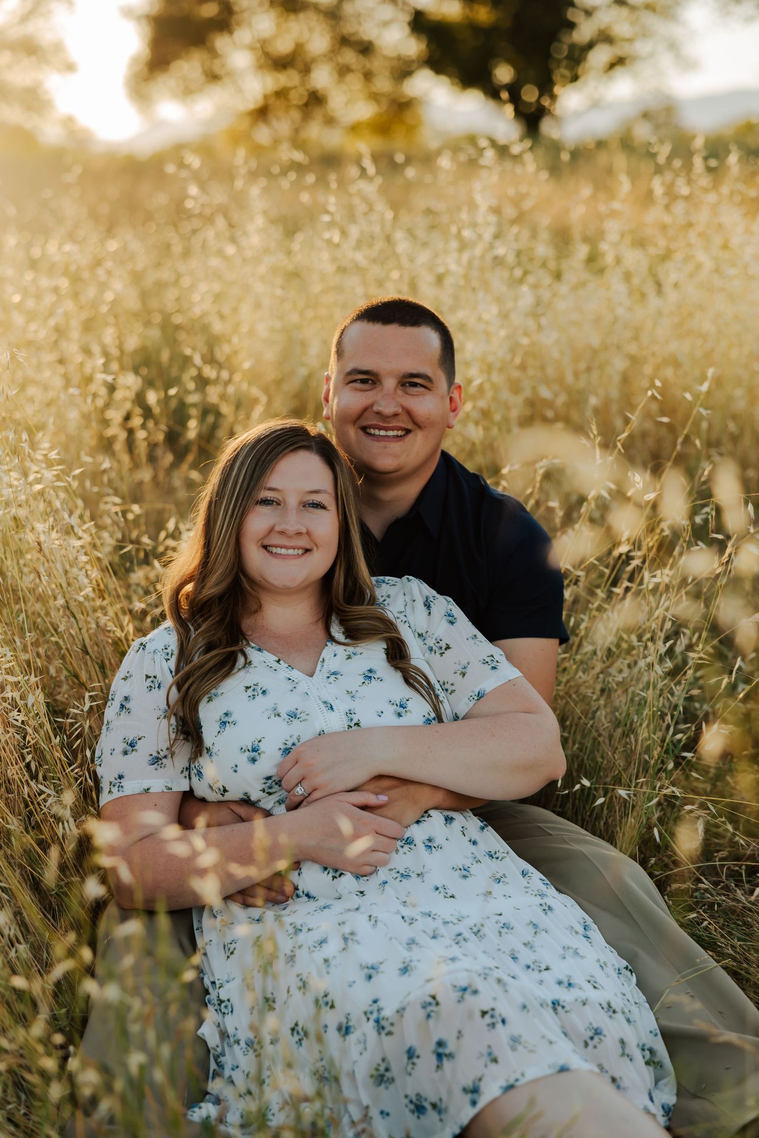 Couple sitting in golden field at sunset, woman in floral dress, man in navy shirt, smiling warmly.