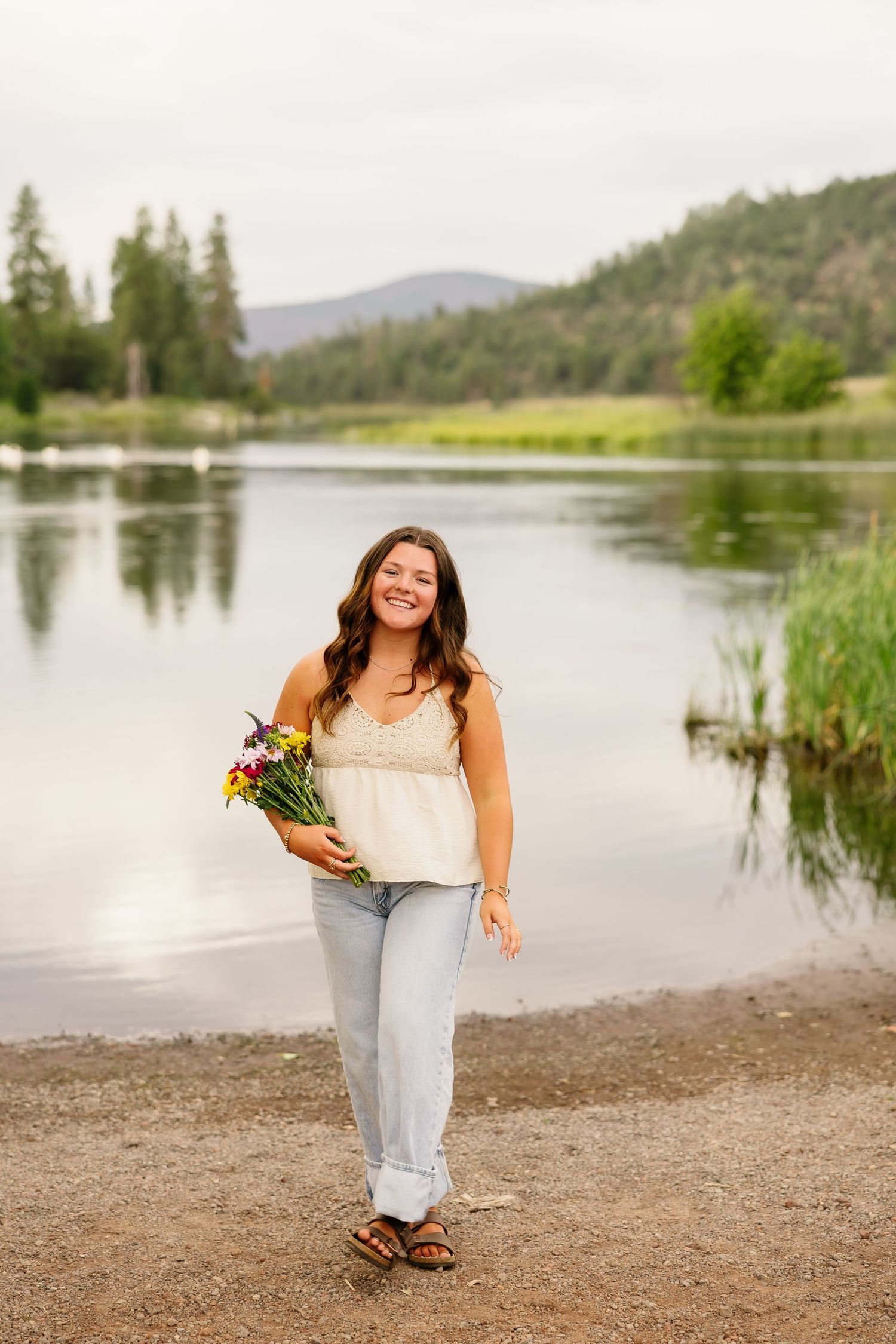 Young woman smiling by a lake holding wildflowers, wearing a white top and jeans with sandals.