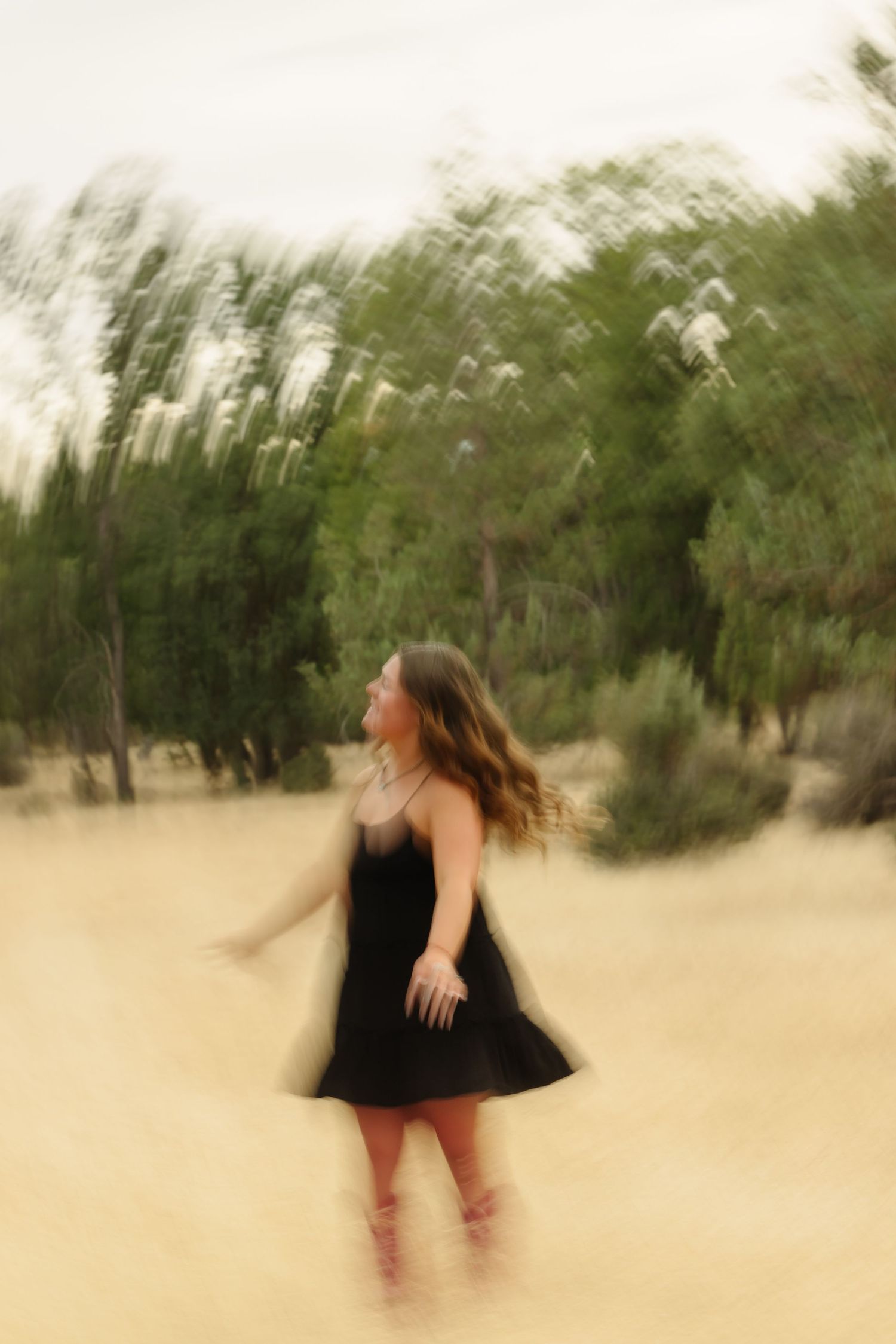 A young woman in a black dress twirls outdoors in a sandy field with green trees, captured with motion blur effect.