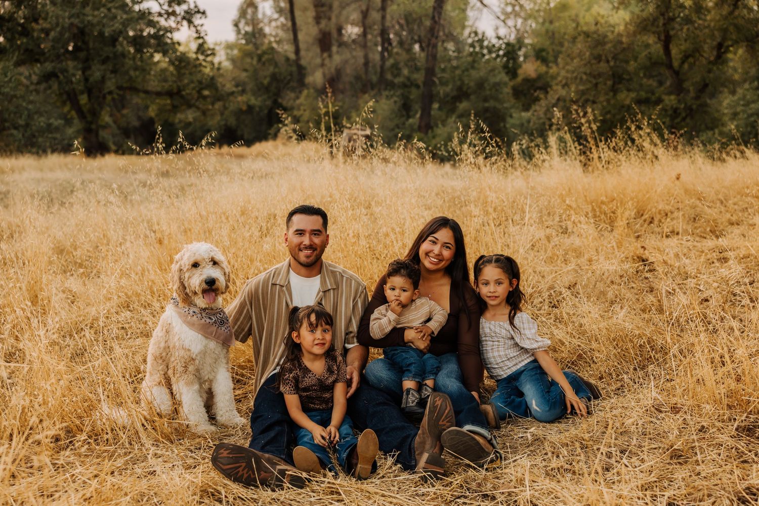 Family and dog gather for casual outdoor portraits in golden grass field.