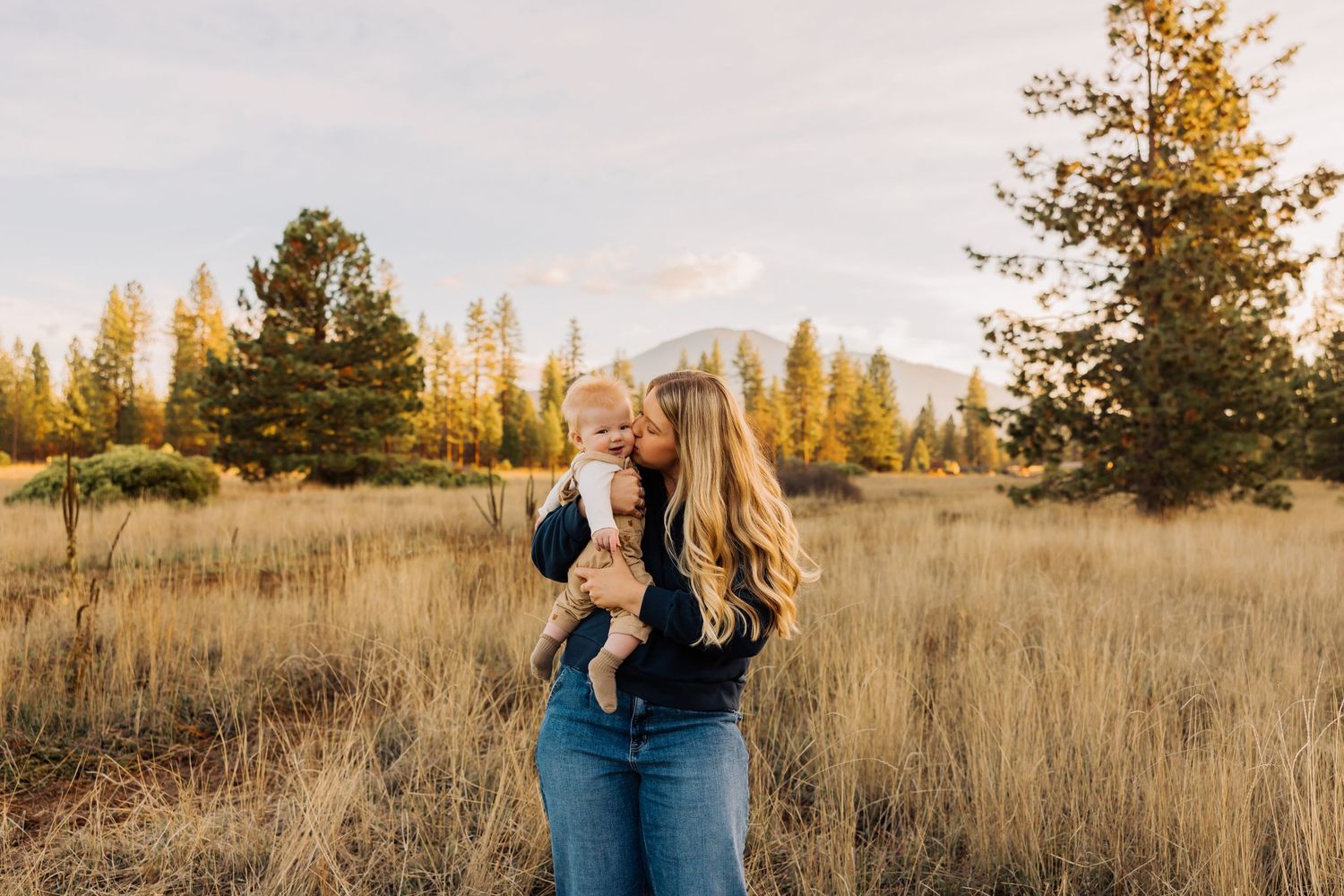 Couple embraces in golden field during sunset with mountains and evergreen trees in background.