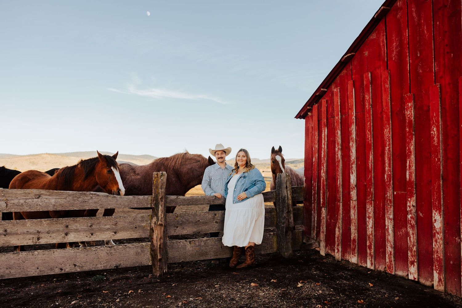 Horses graze near a weathered red barn as the sun sets over the prairie.