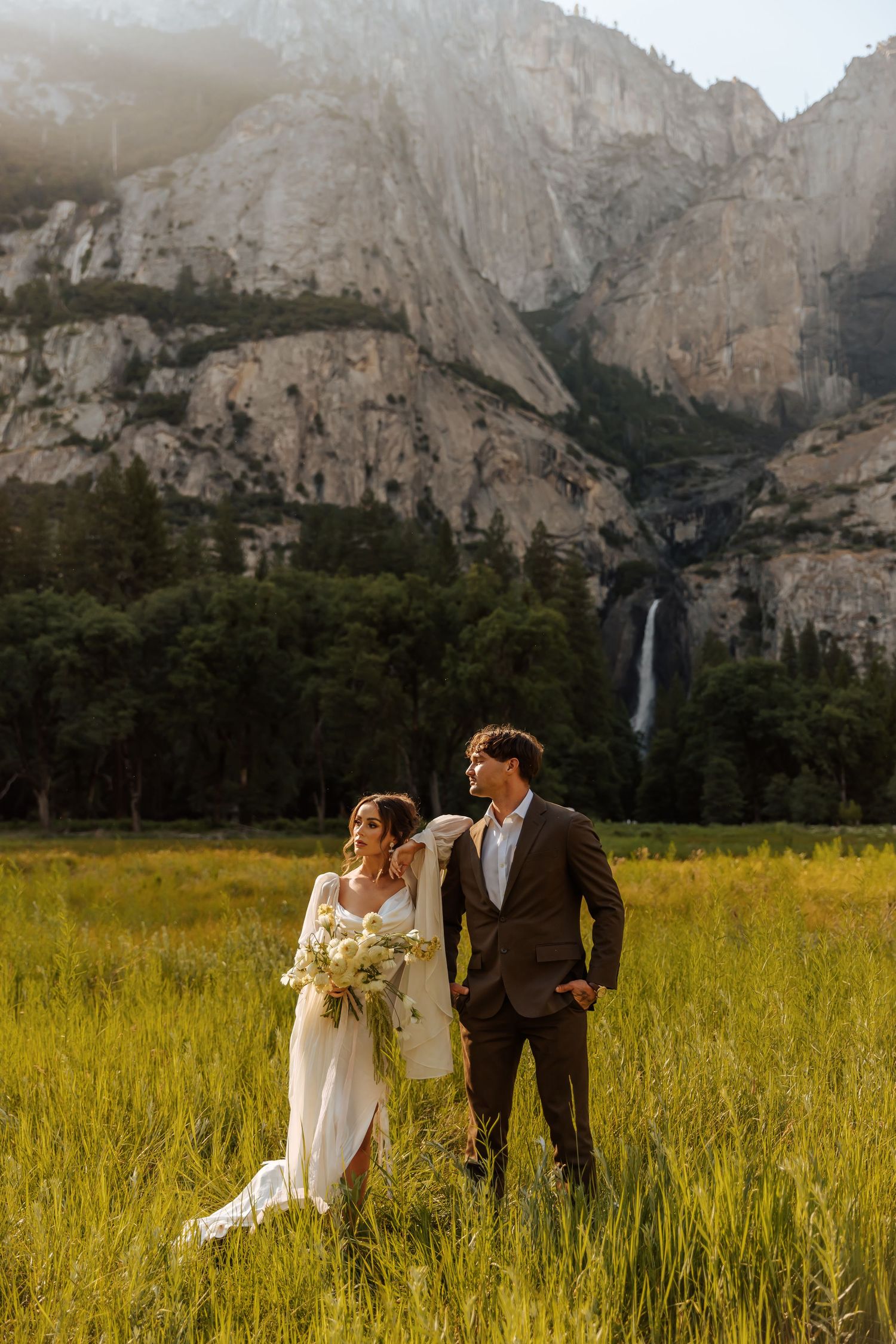 A couple stands in a golden meadow with dramatic granite cliffs and cascading waterfalls of Yosemite Valley in the background.