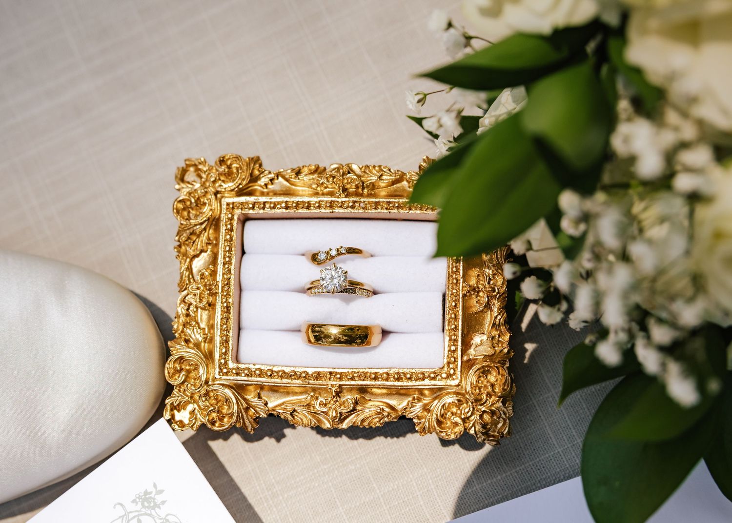 Gold ornate frame box displaying wedding bands and diamond engagement ring beside white flowers on elegant surface.