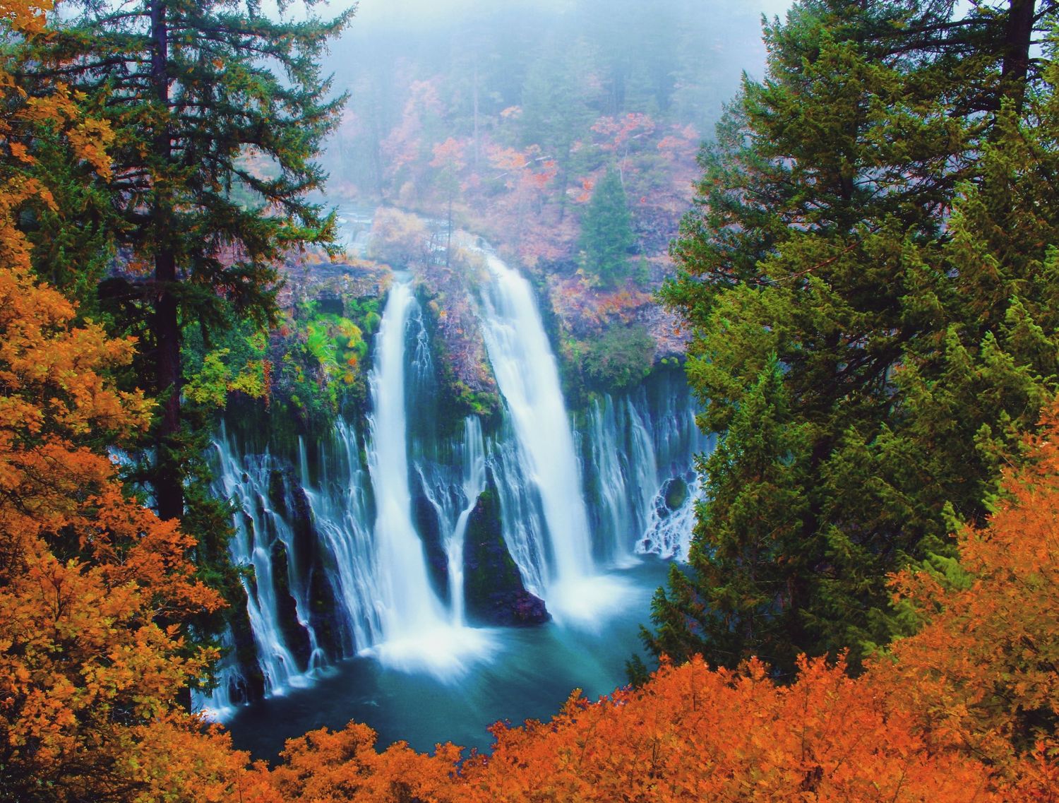 Stunning autumn waterfall cascades through vibrant orange foliage and evergreen trees into a turquoise pool below.