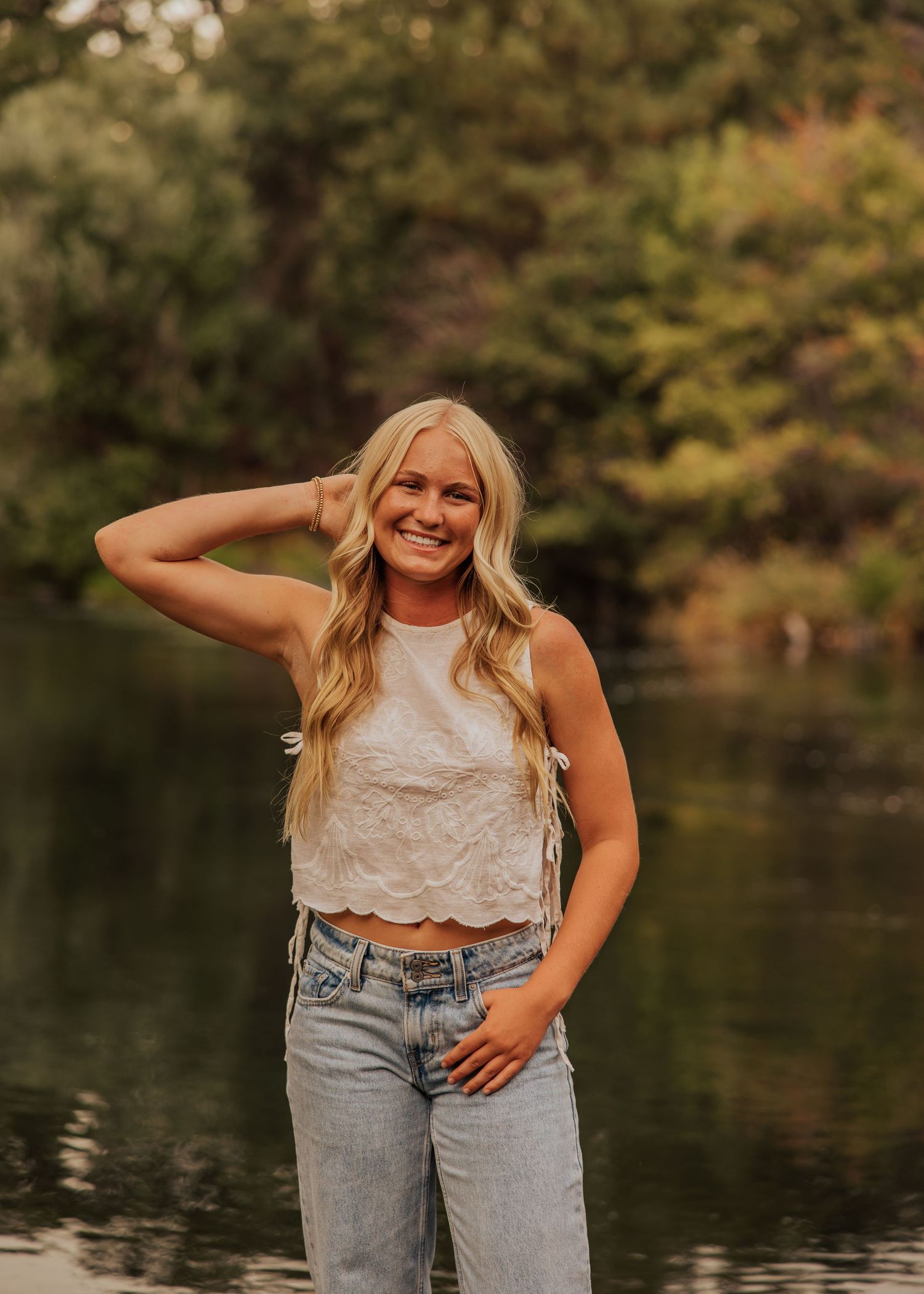 Person posing by lake wearing white lace top and jeans in natural setting.