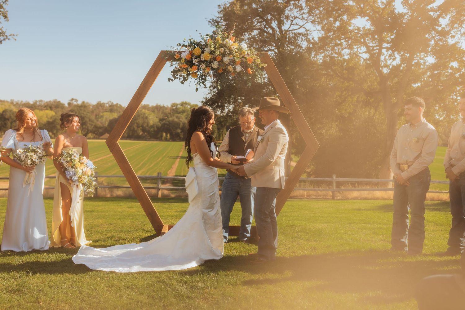 Golden hour wedding ceremony setup with wooden arch and guests on grassy field.
