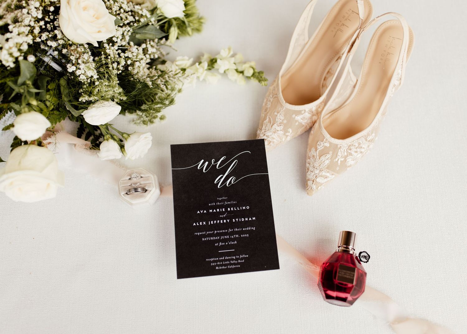 Elegant wedding flat lay with black ‘we do’ invitation, lace bridal heels, white floral bouquet, and red perfume bottle.