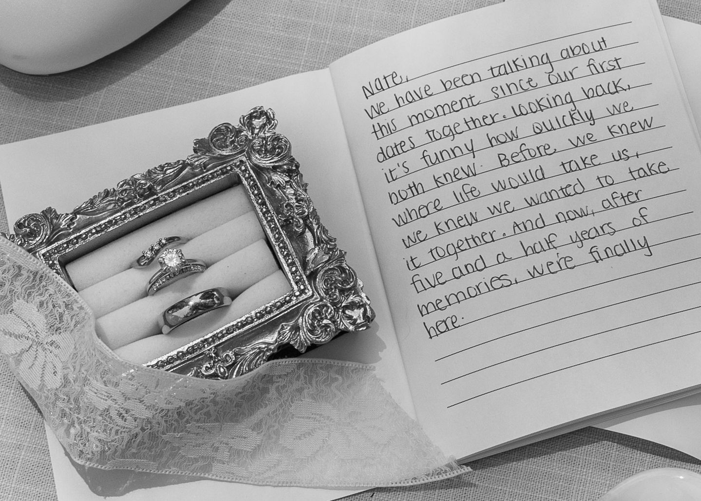 Black and white photo of wedding rings in ornate frame beside open journal with handwritten love letter.