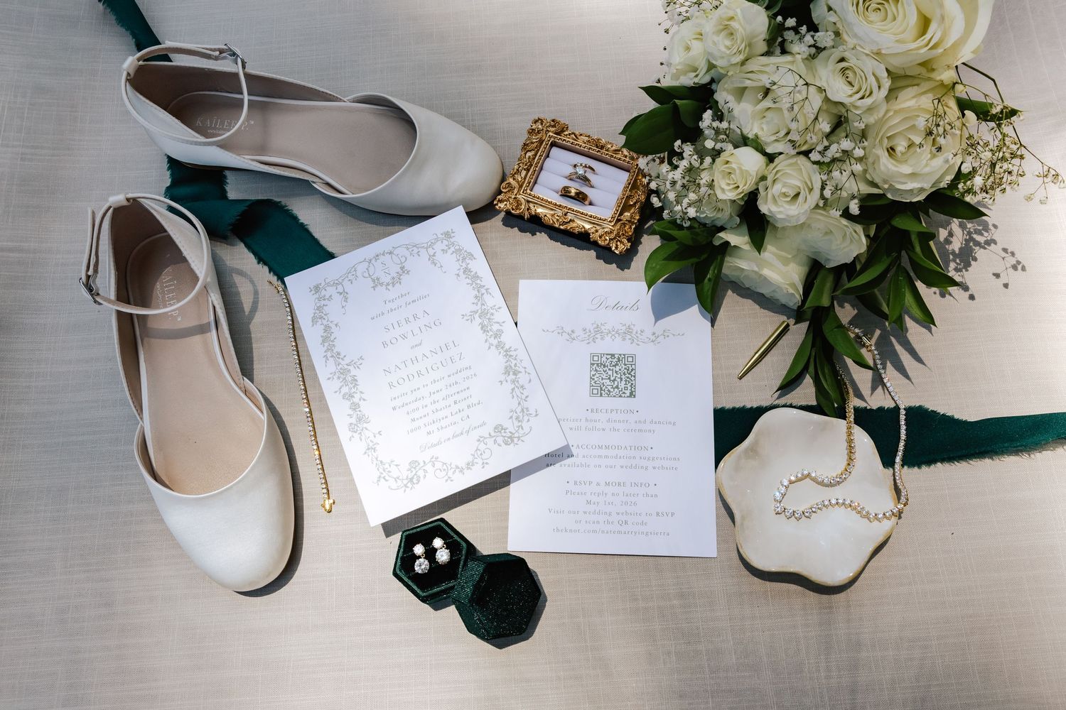 Elegant wedding flat lay featuring ivory bridal shoes, green velvet ribbon, white floral bouquet, invitations, and pearl jewelry.