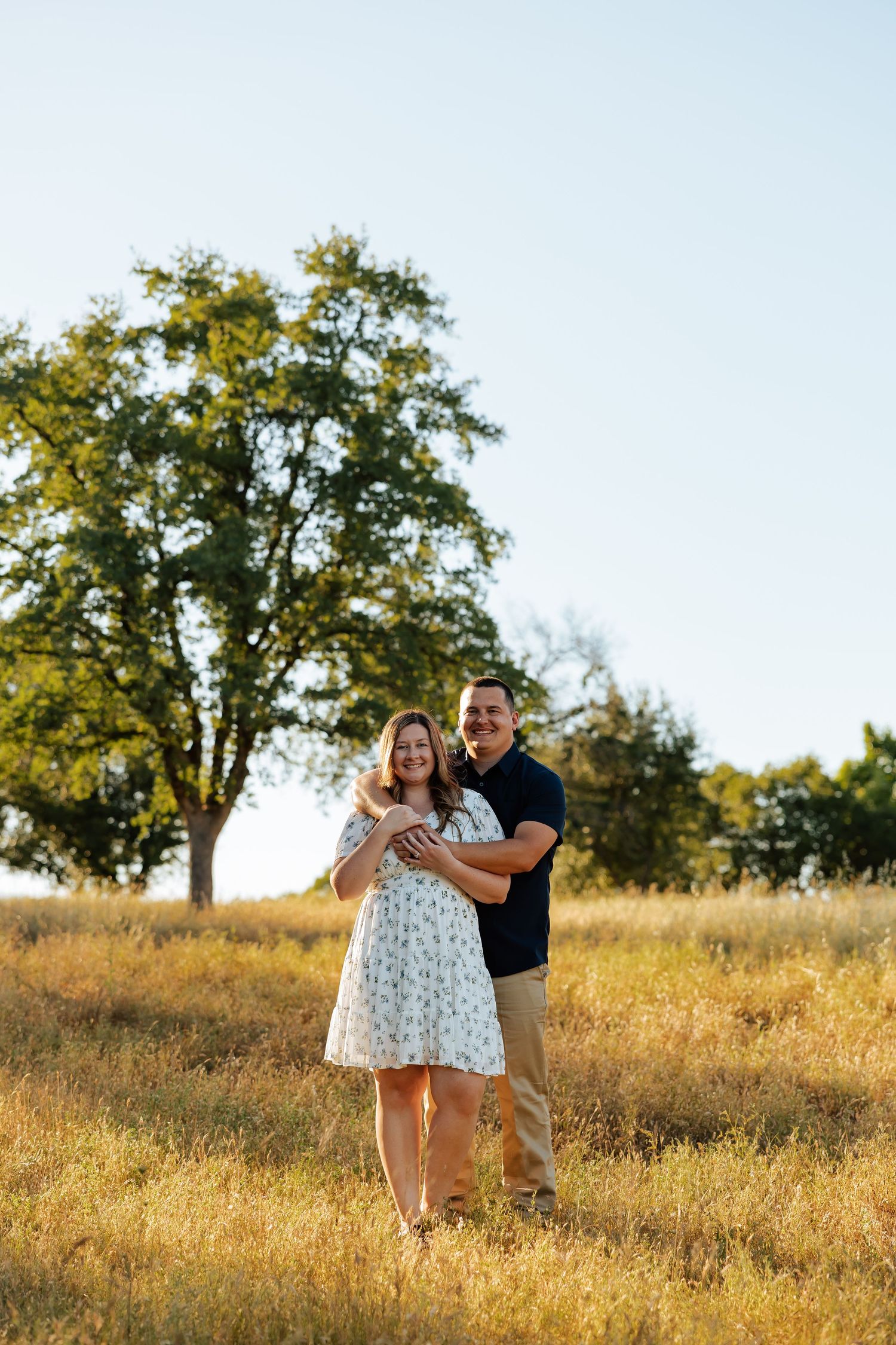 A couple embraces in a sunny golden field with a large green tree behind them on a clear day.