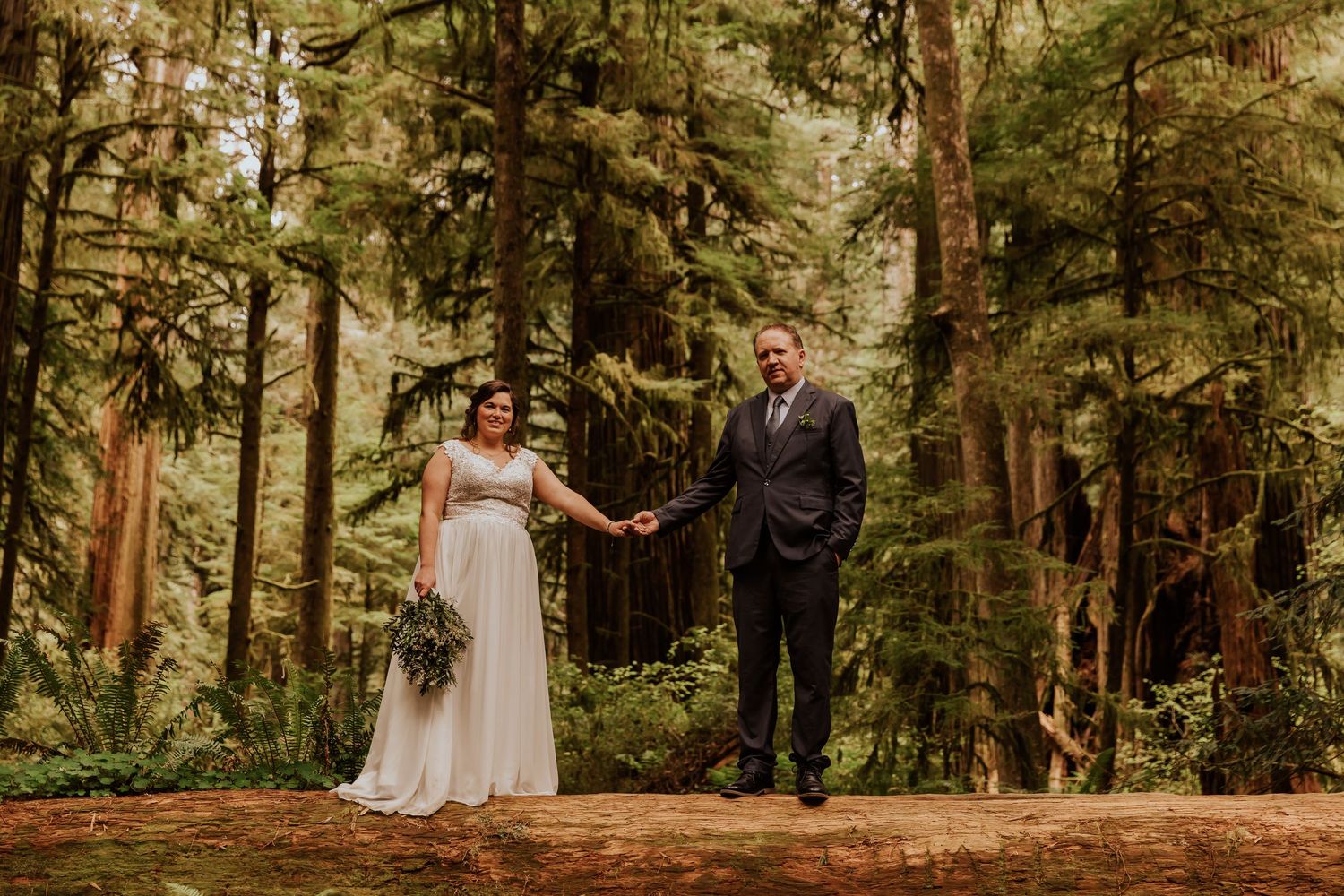 A wide forest wedding scene showing guests and couple amid majestic redwood trees.