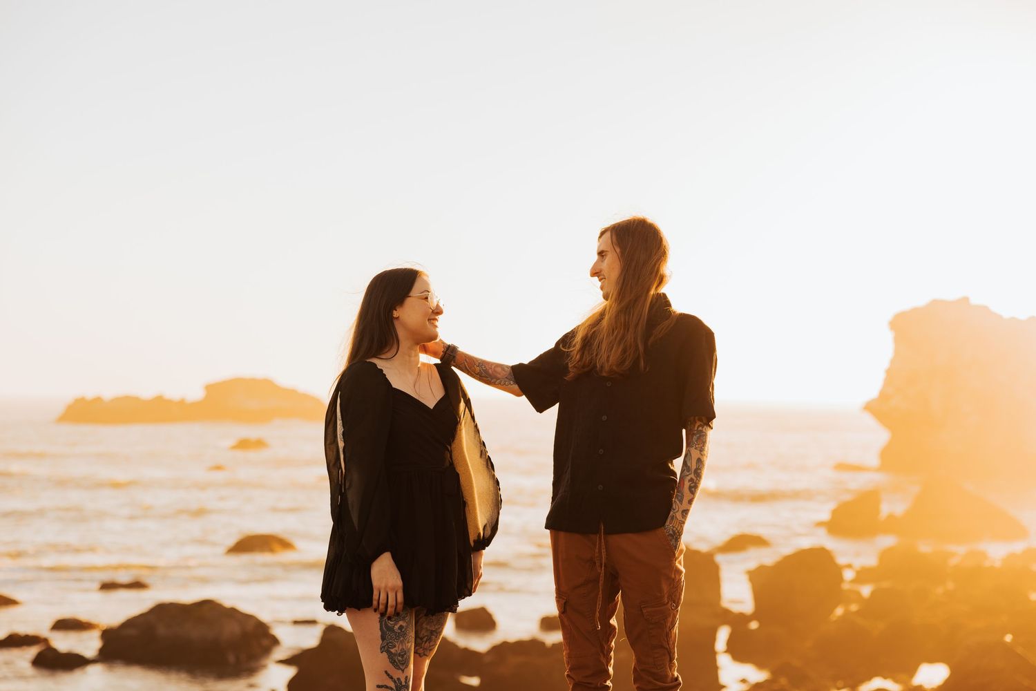 Two people share laughter on coastal rocks during a golden sunset.
