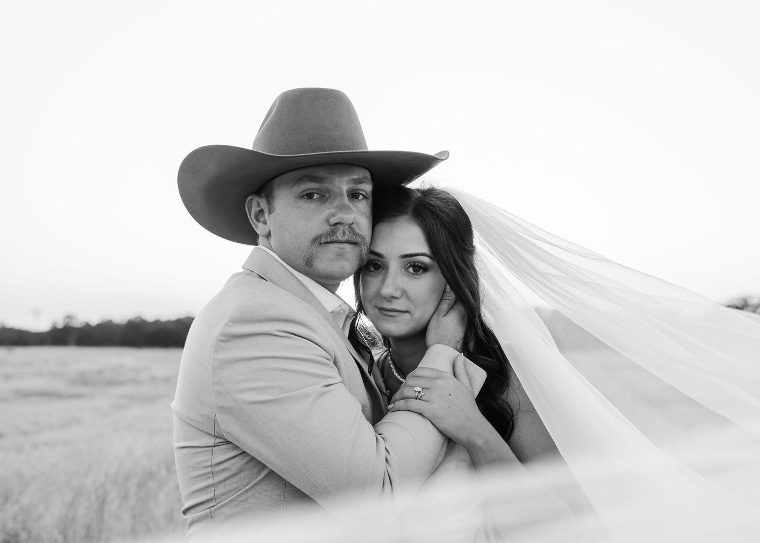 Black and white wedding photo of bride and groom embracing in a field, groom wearing cowboy hat, bride in veil.