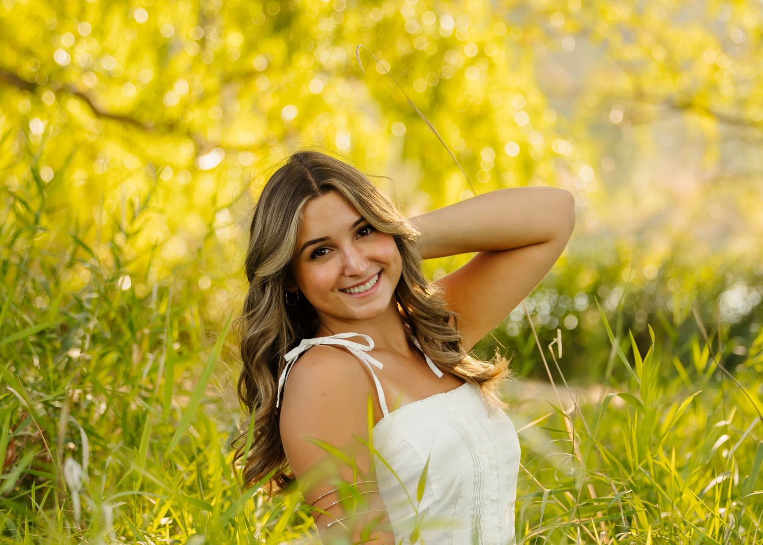 Young woman in white dress smiling in lush green grass with vibrant yellow bokeh trees in background.
