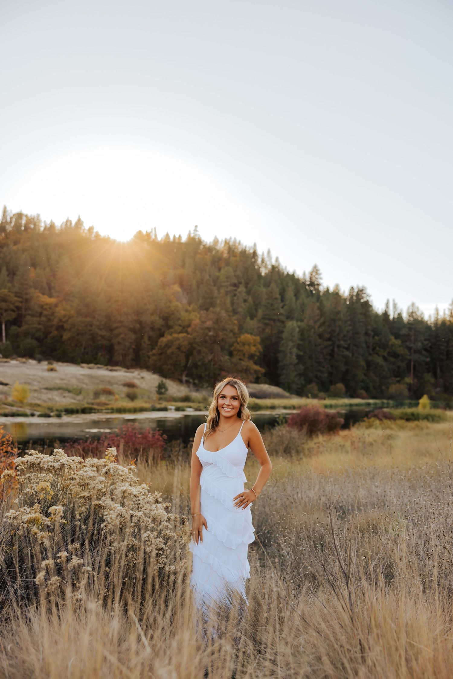 Woman in white dress standing in golden field at sunset with mountain and lake backdrop.