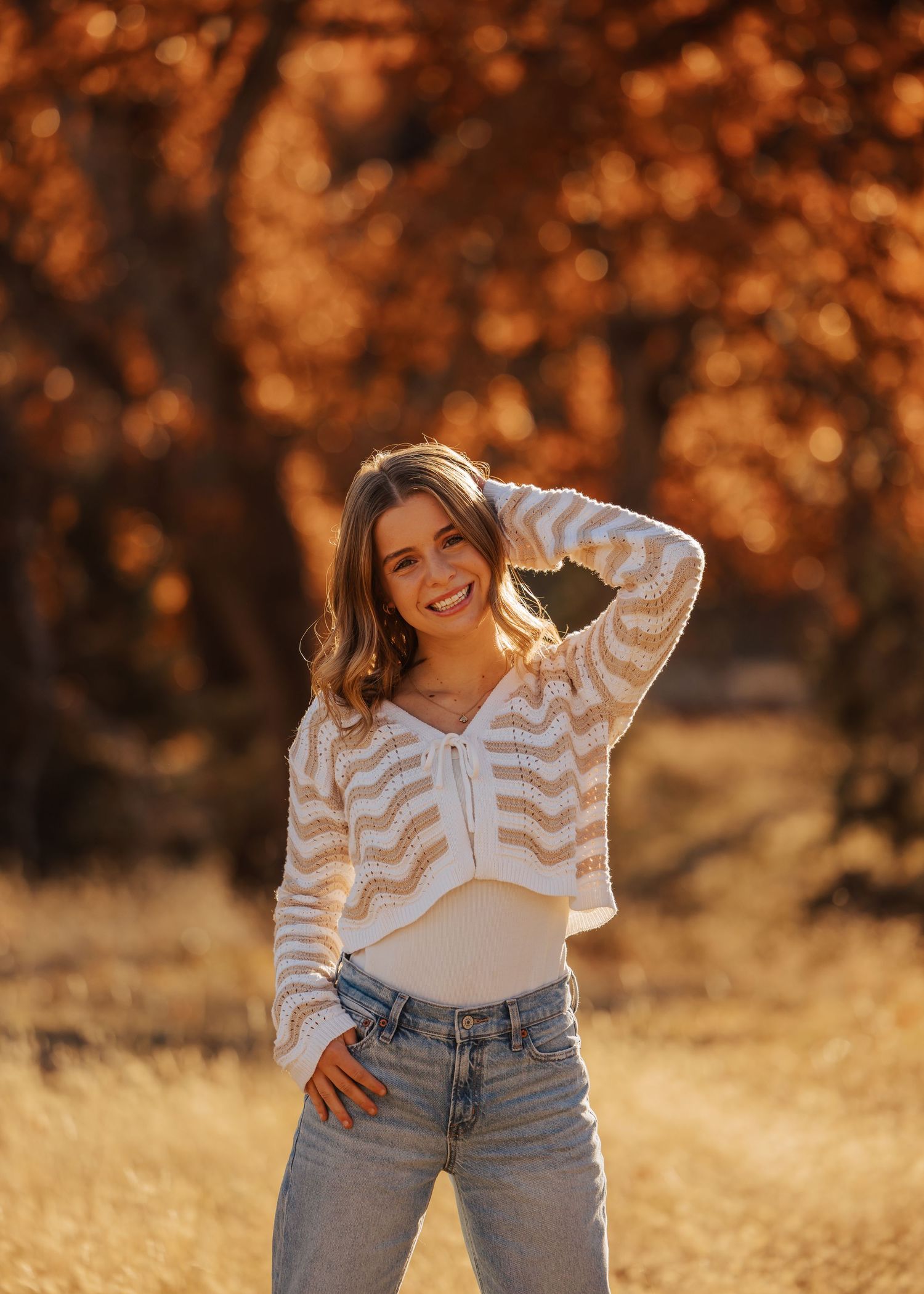 Someone wearing a striped sweater and jeans poses playfully in an autumn field with golden lighting.