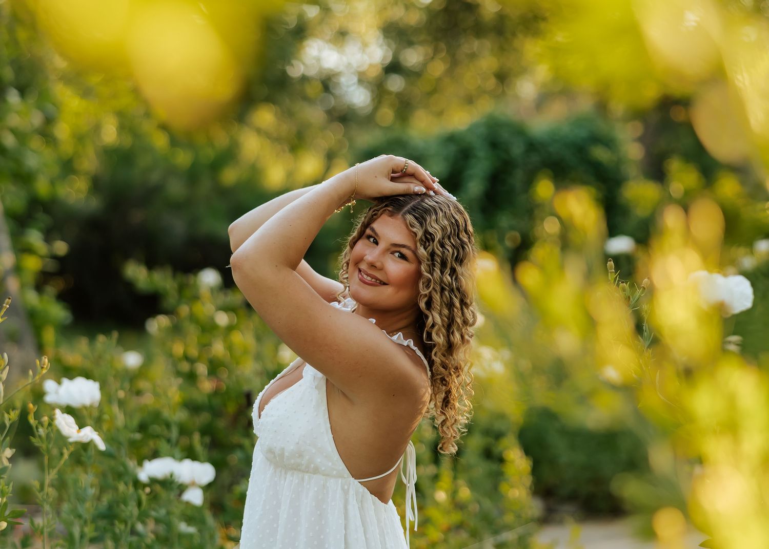 Young woman in white dress posing gracefully among yellow and white flowers in a sunlit garden.