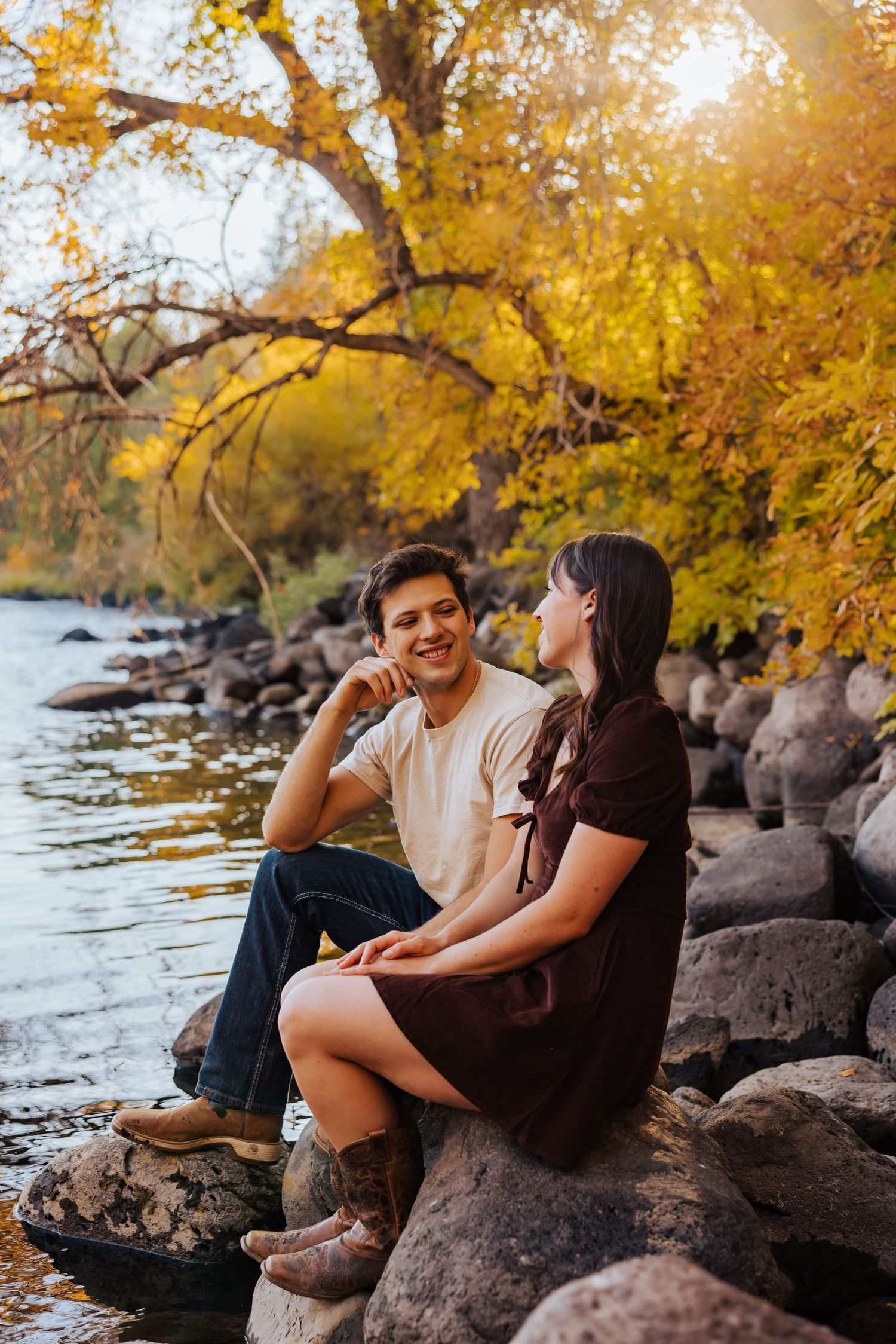 Couple relaxes on rocky riverbank beneath golden autumn trees.