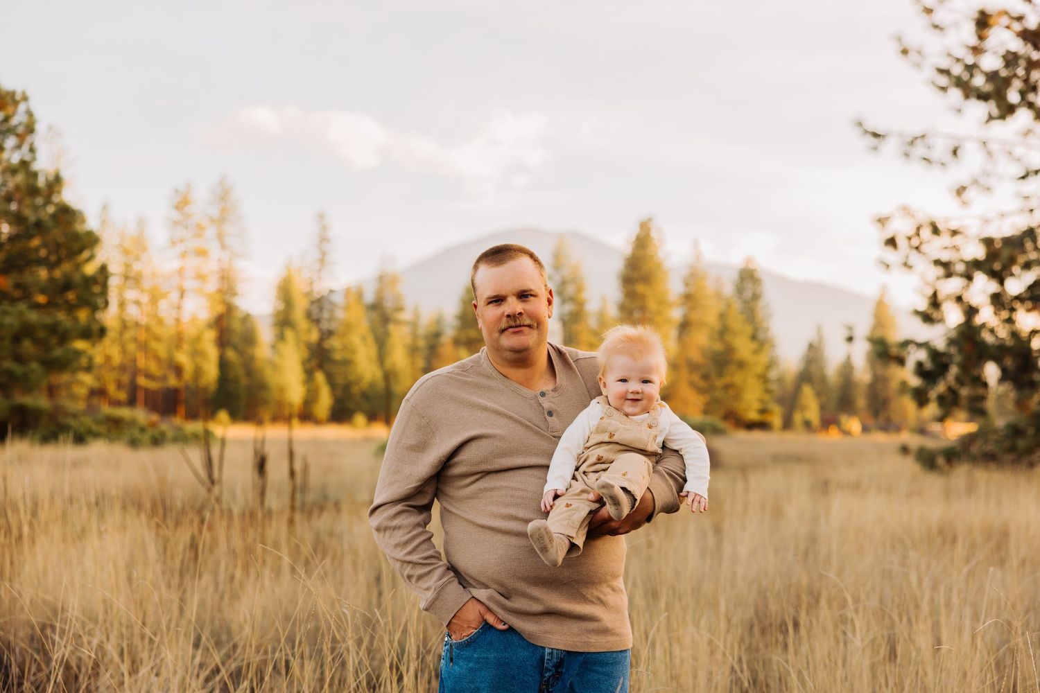 A father holds his baby while standing in a golden mountain meadow at sunset.