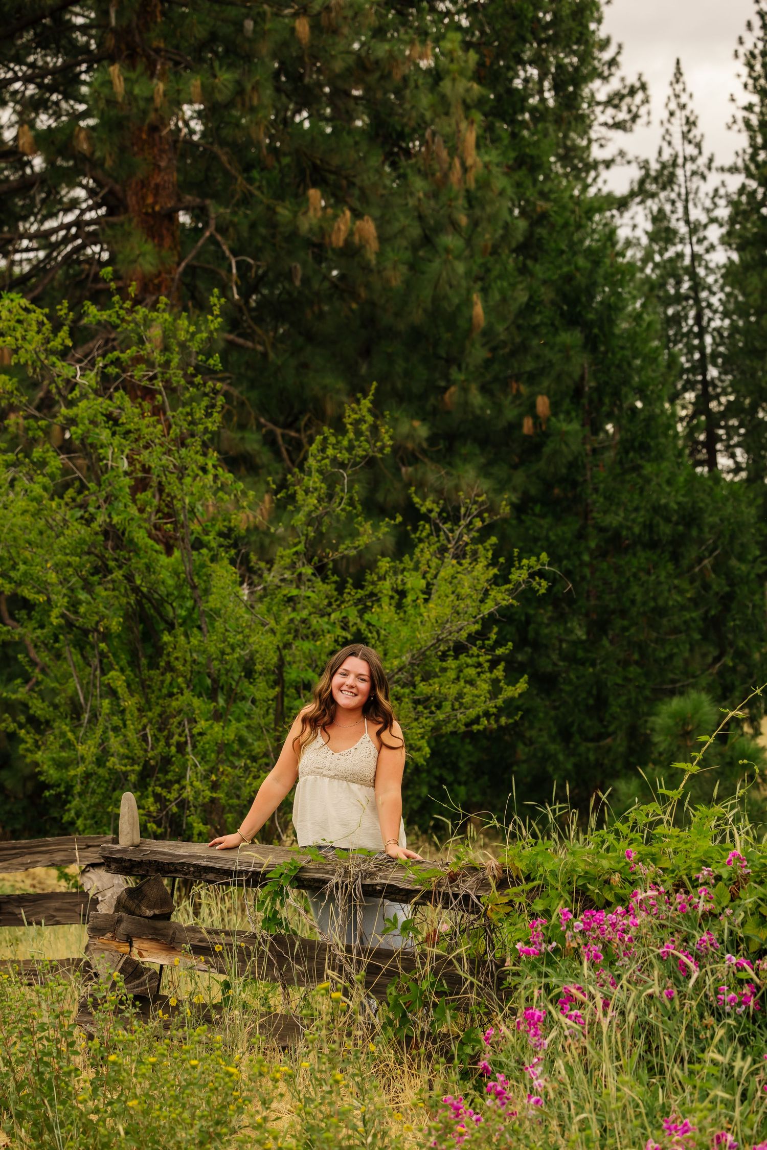 Woman in white dress smiling outdoors near rustic fence with wildflowers and tall evergreen trees in background.