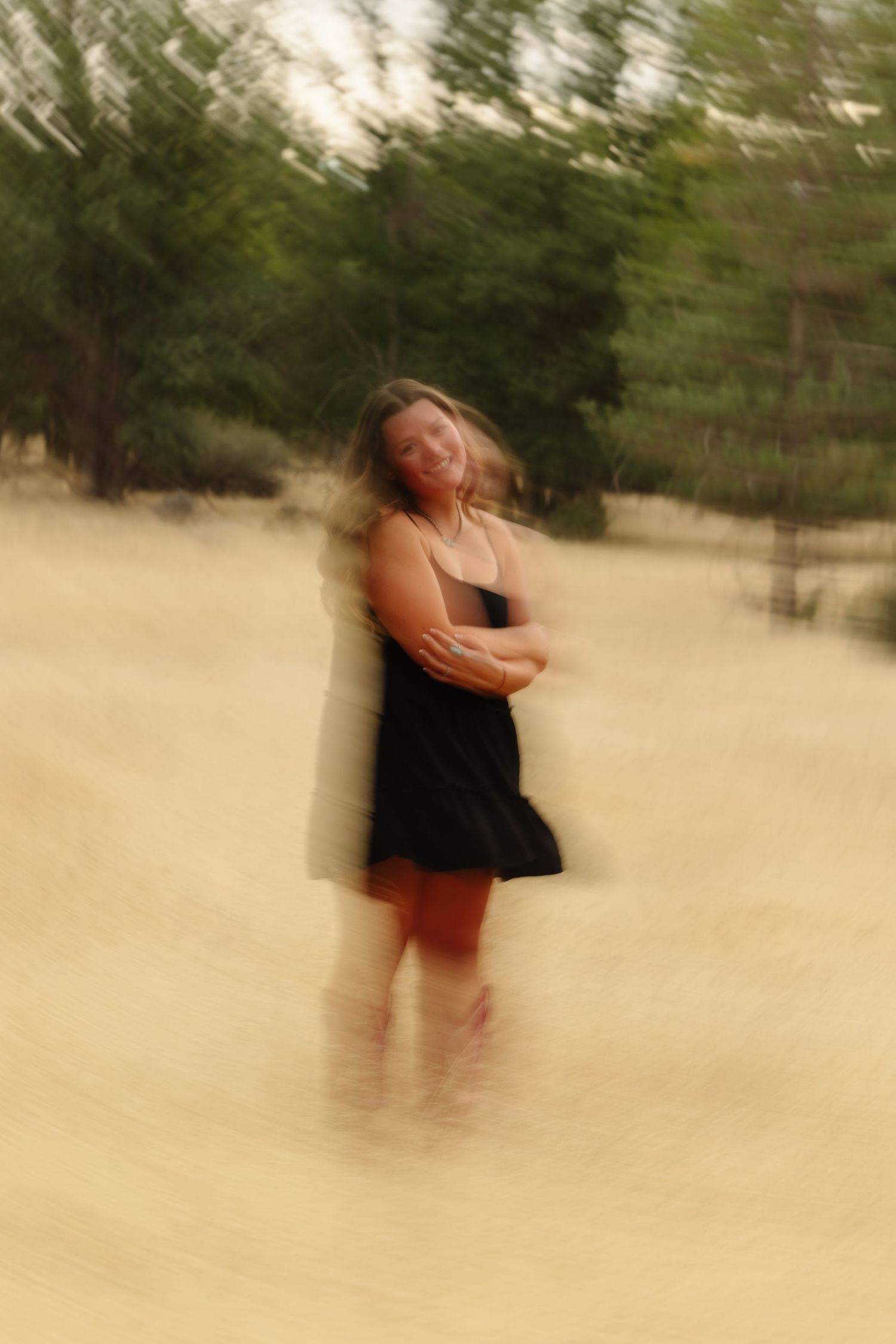 A woman in a black dress stands on sandy ground with trees behind her, captured with a motion blur artistic effect.