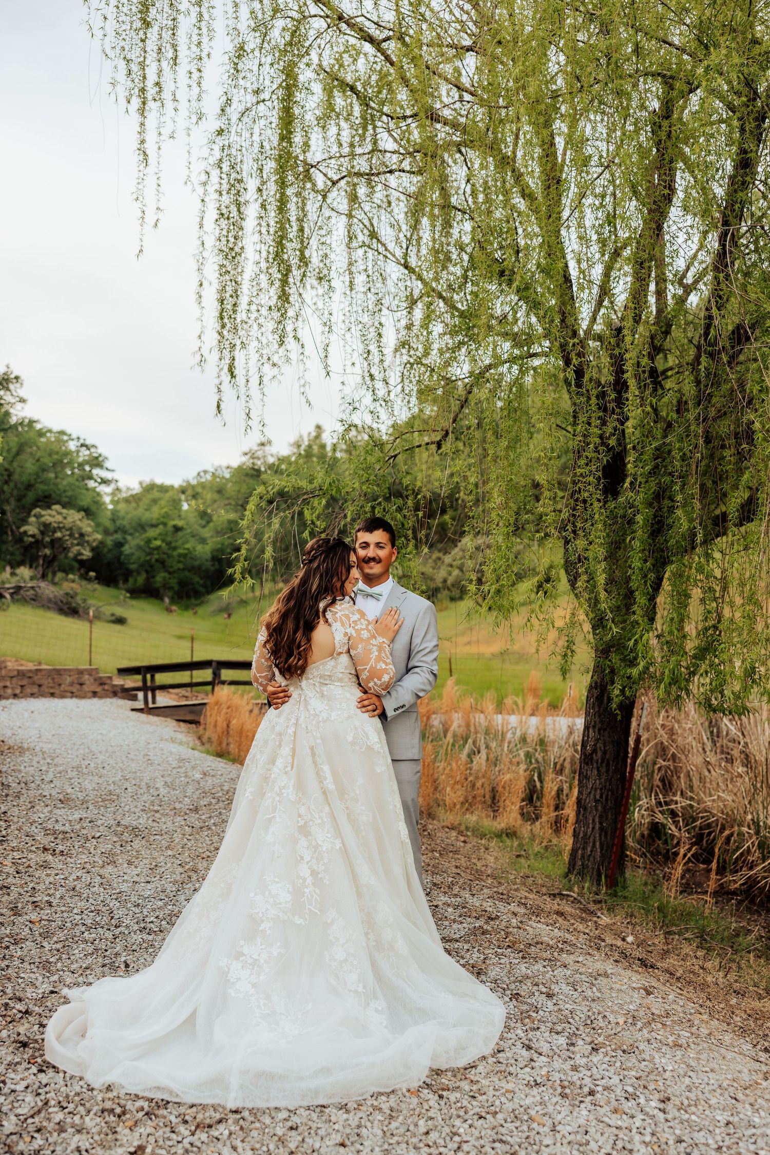 Bride and groom pose on gravel path beside weeping willow tree in lush green outdoor wedding setting.