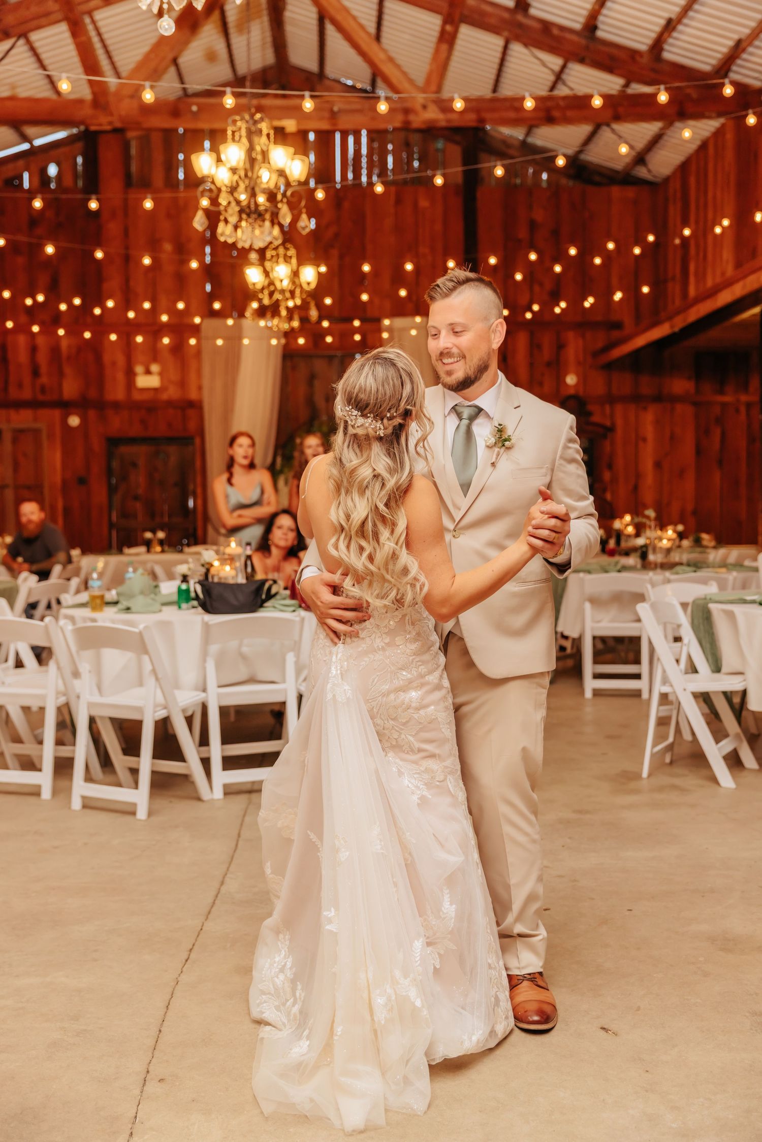 Newlyweds share first dance in rustic barn reception venue with string lights and chandeliers.