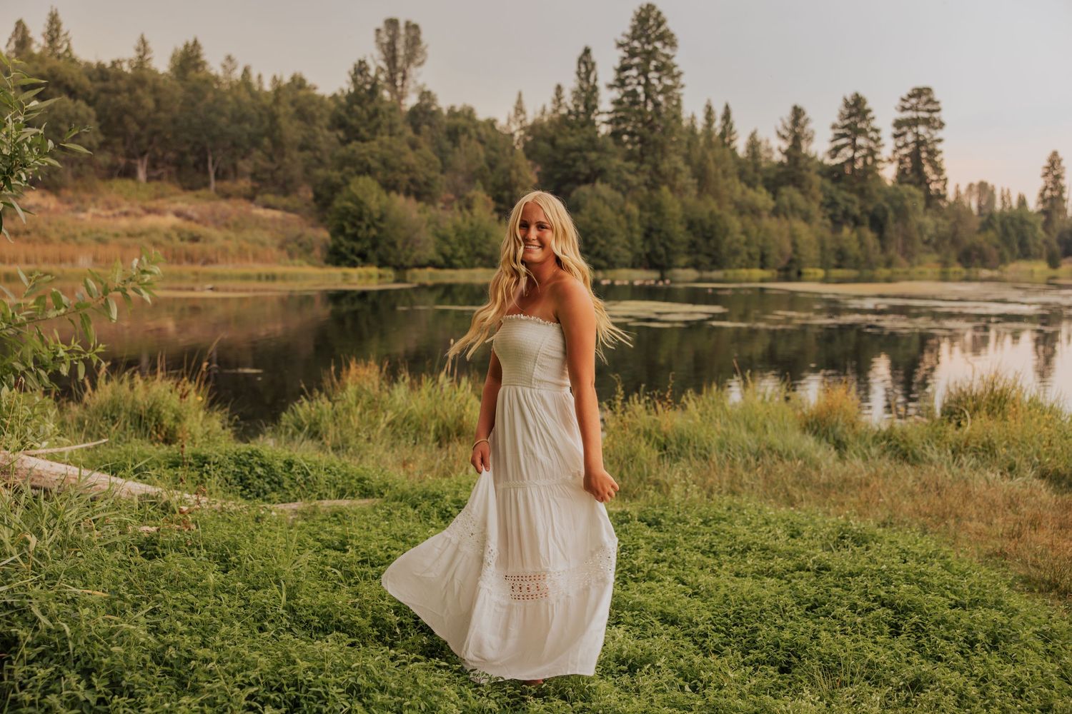 Person in white dress standing near lake with evergreen trees in background during golden hour.