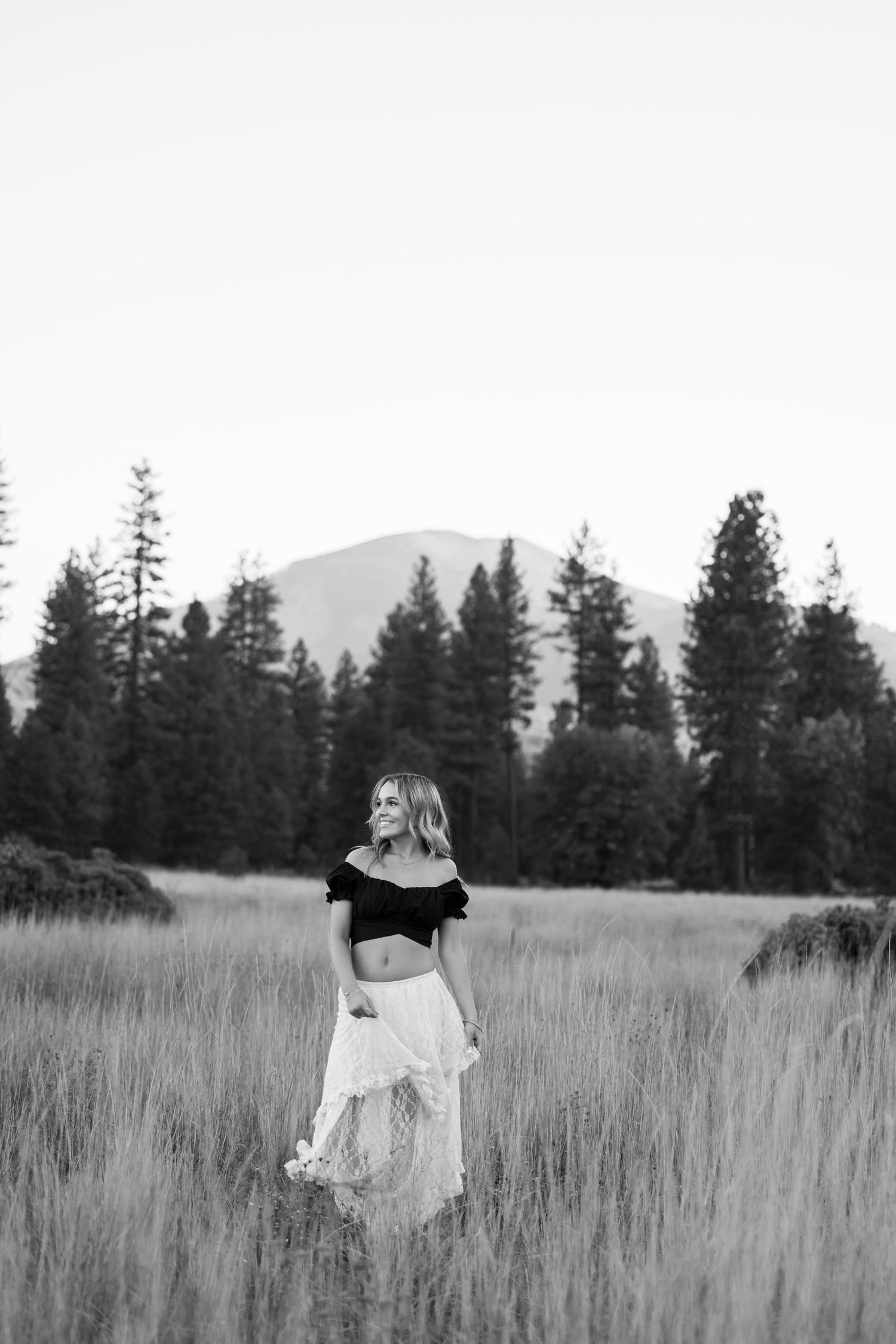 Figure in crop top and flowing skirt stands in mountain meadow with evergreen forest backdrop.