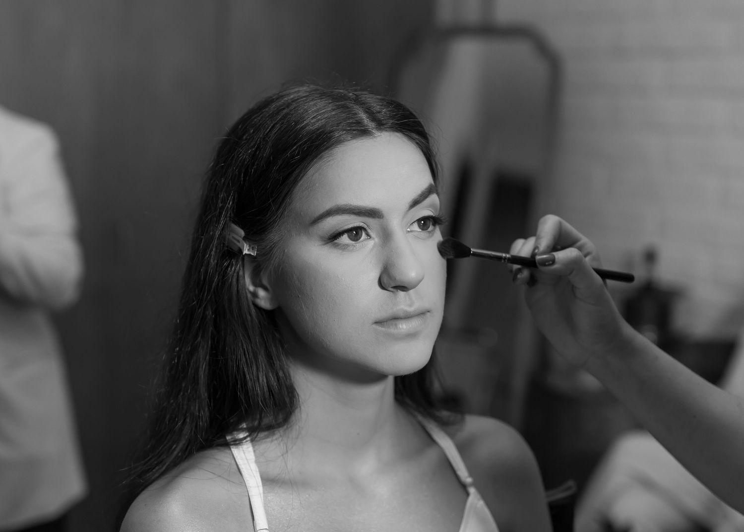 Black and white photo of a young woman getting makeup applied with a brush by a professional artist.