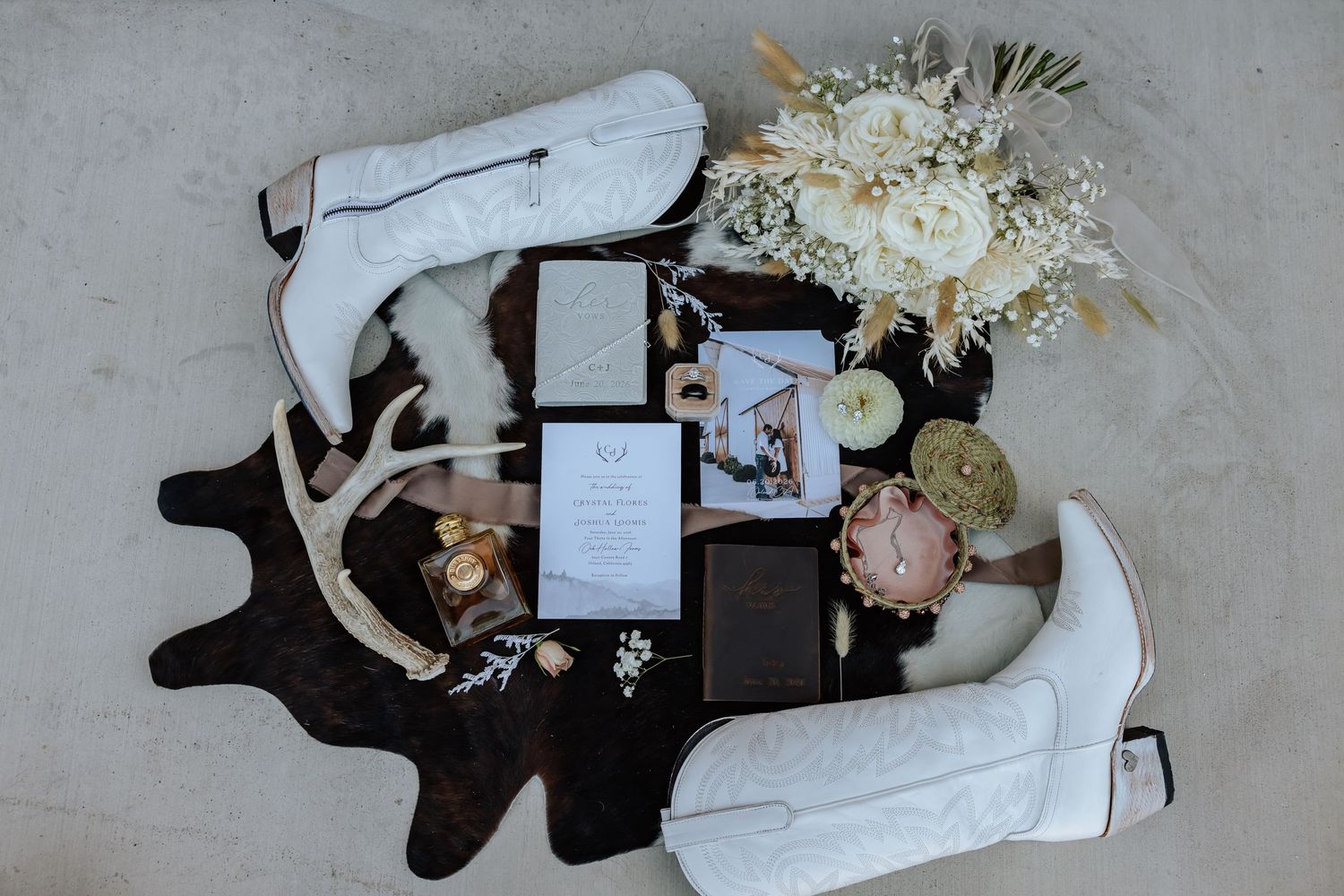 White cowboy boots, wedding invitations, floral bouquet, and antlers styled on cowhide rug for rustic western wedding flat lay.