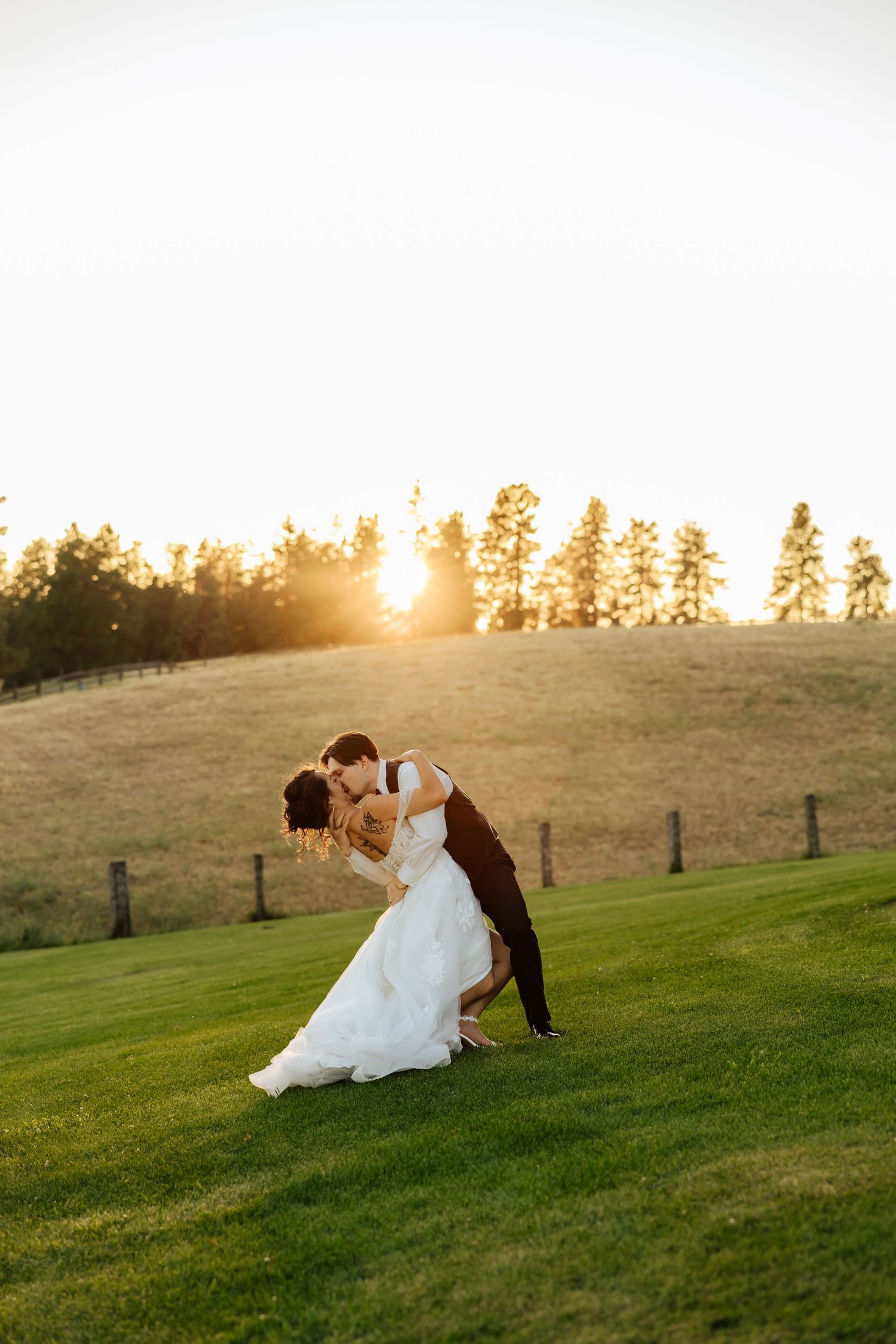 Groom dips bride for a kiss at golden sunset on lush green hillside.