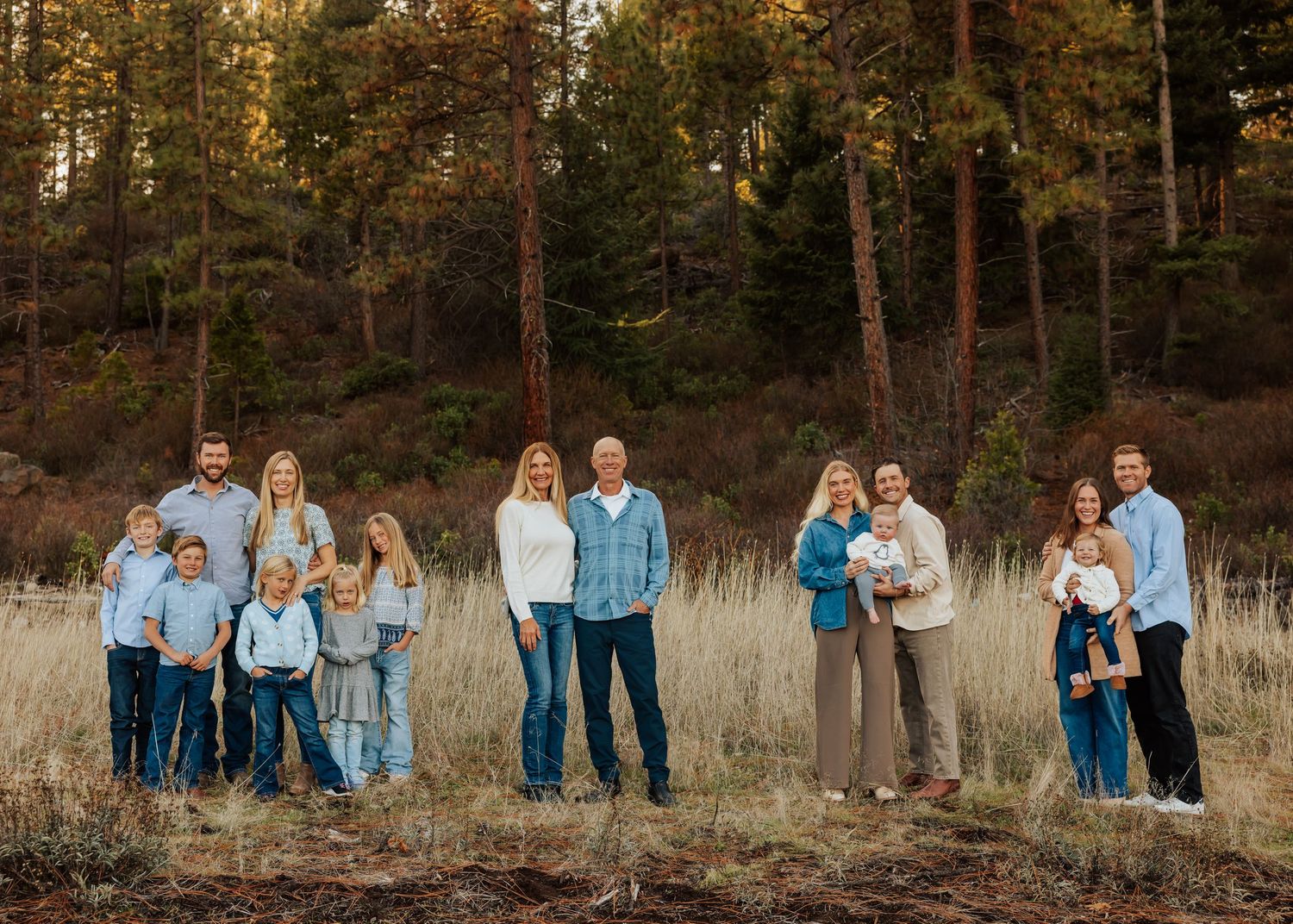 Extended family portrait in casual denim and blue tones standing in a golden field with pine trees.