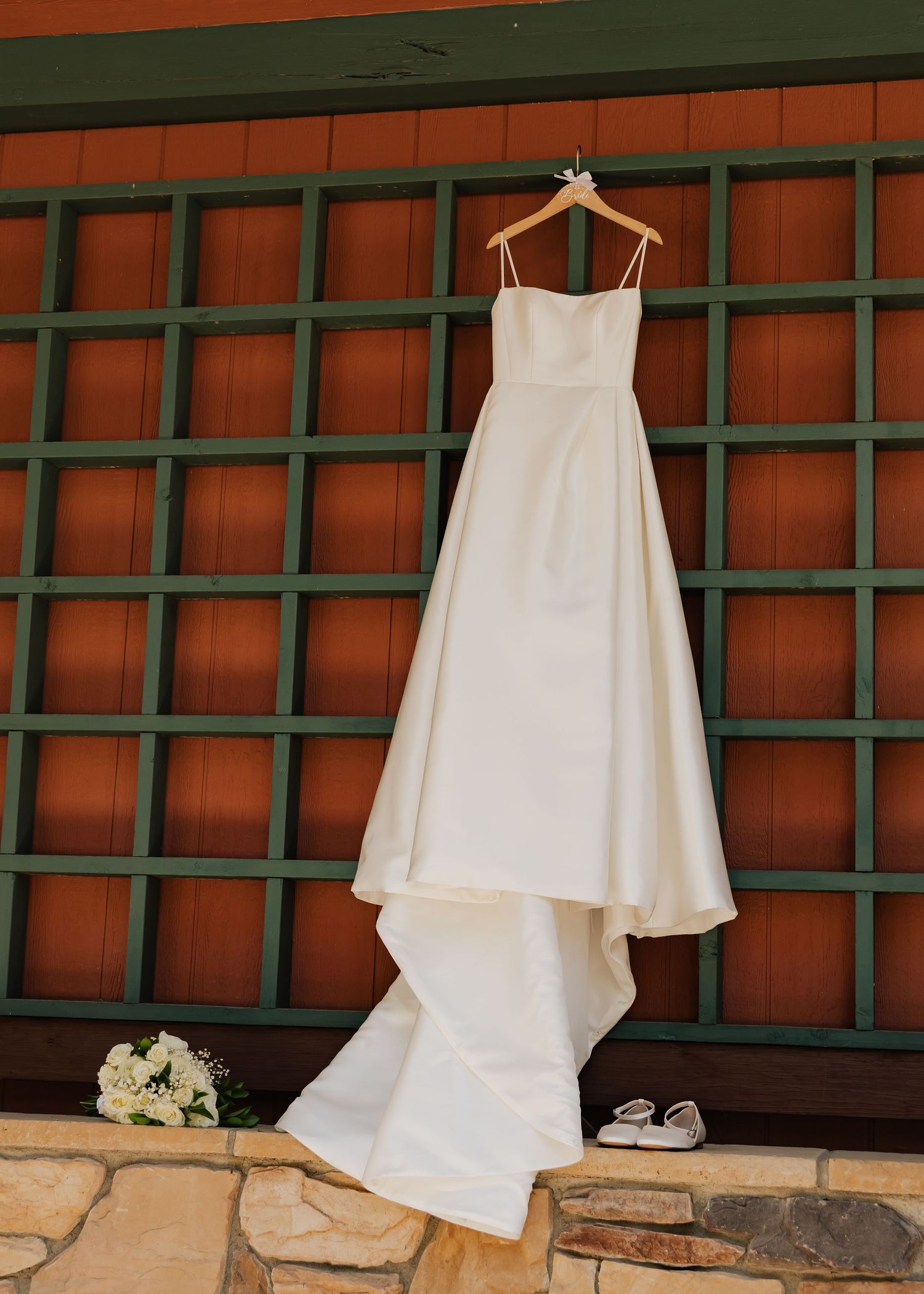 Elegant white spaghetti strap wedding gown hanging on wooden lattice wall with bridal bouquet and shoes below.