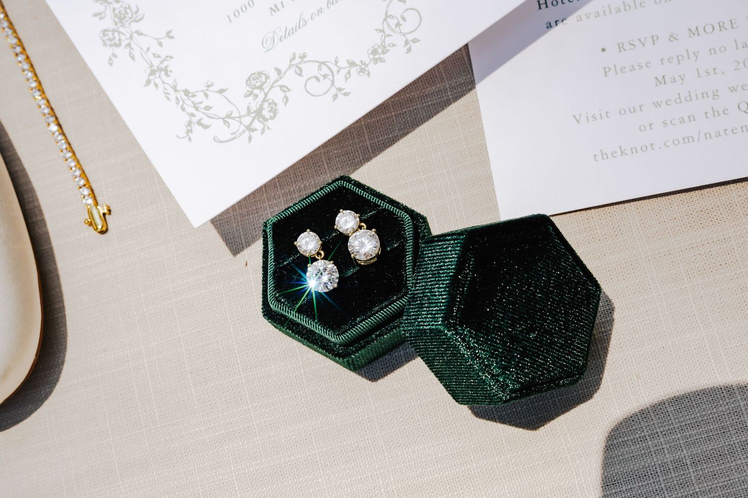 Green velvet hexagon jewelry box displaying sparkling diamond stud earrings beside a wedding invitation and gold necklace.