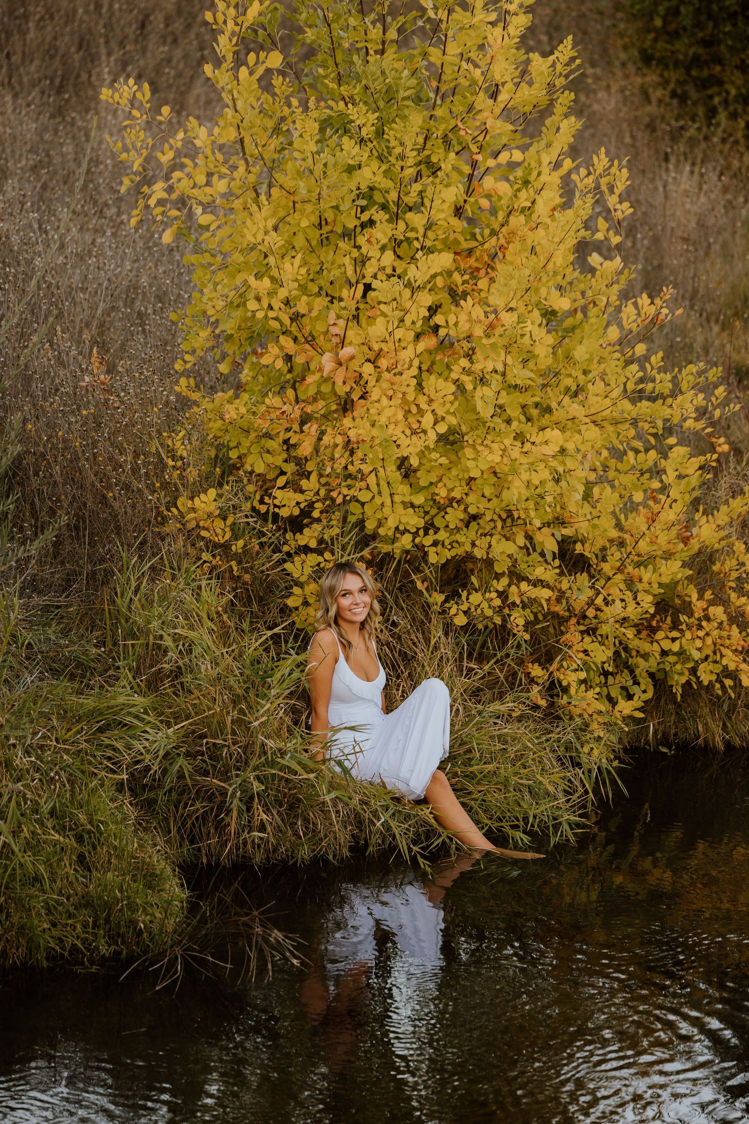 Woman in white dress perches on riverbank beside vibrant yellow fall foliage.