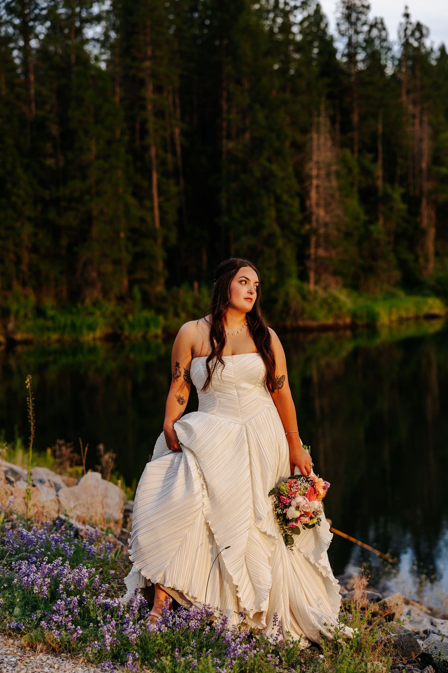 Bride in strapless white gown holds bouquet by a forest lake at golden hour.