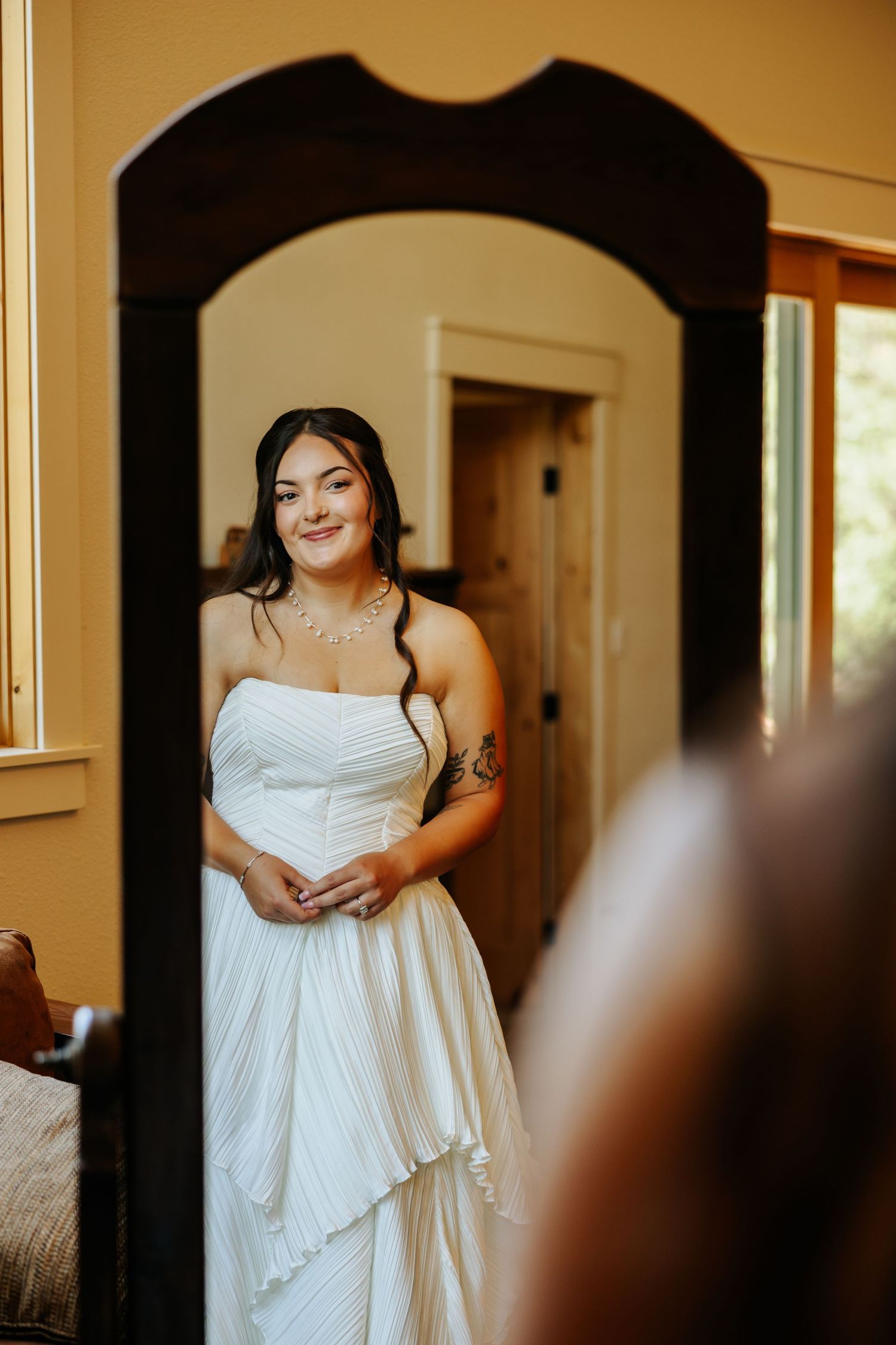 Bride in strapless white gown smiling at her reflection in an ornate dark wood mirror before her wedding.