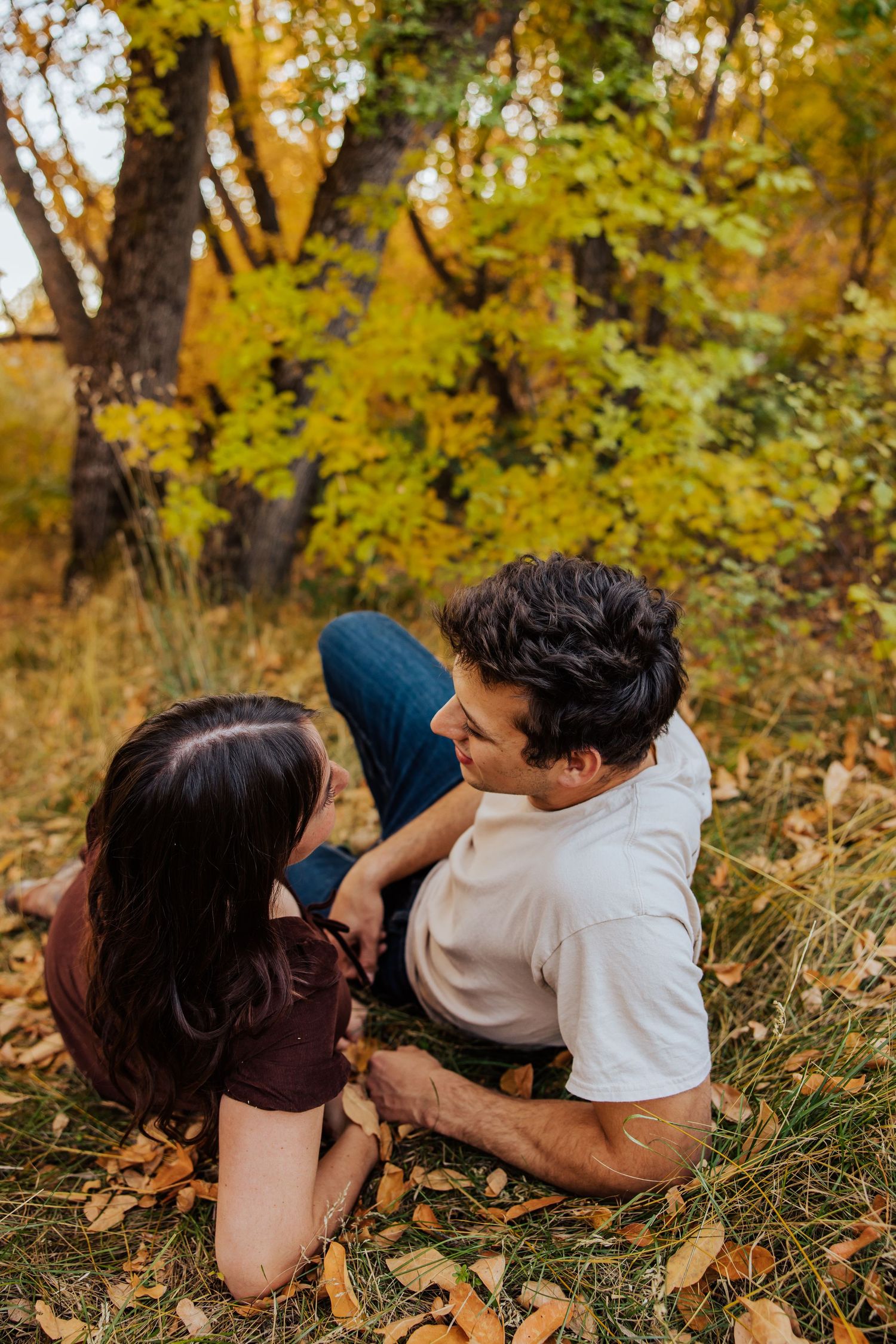 Couple lying together playfully among fallen leaves in a sunlit autumn forest.