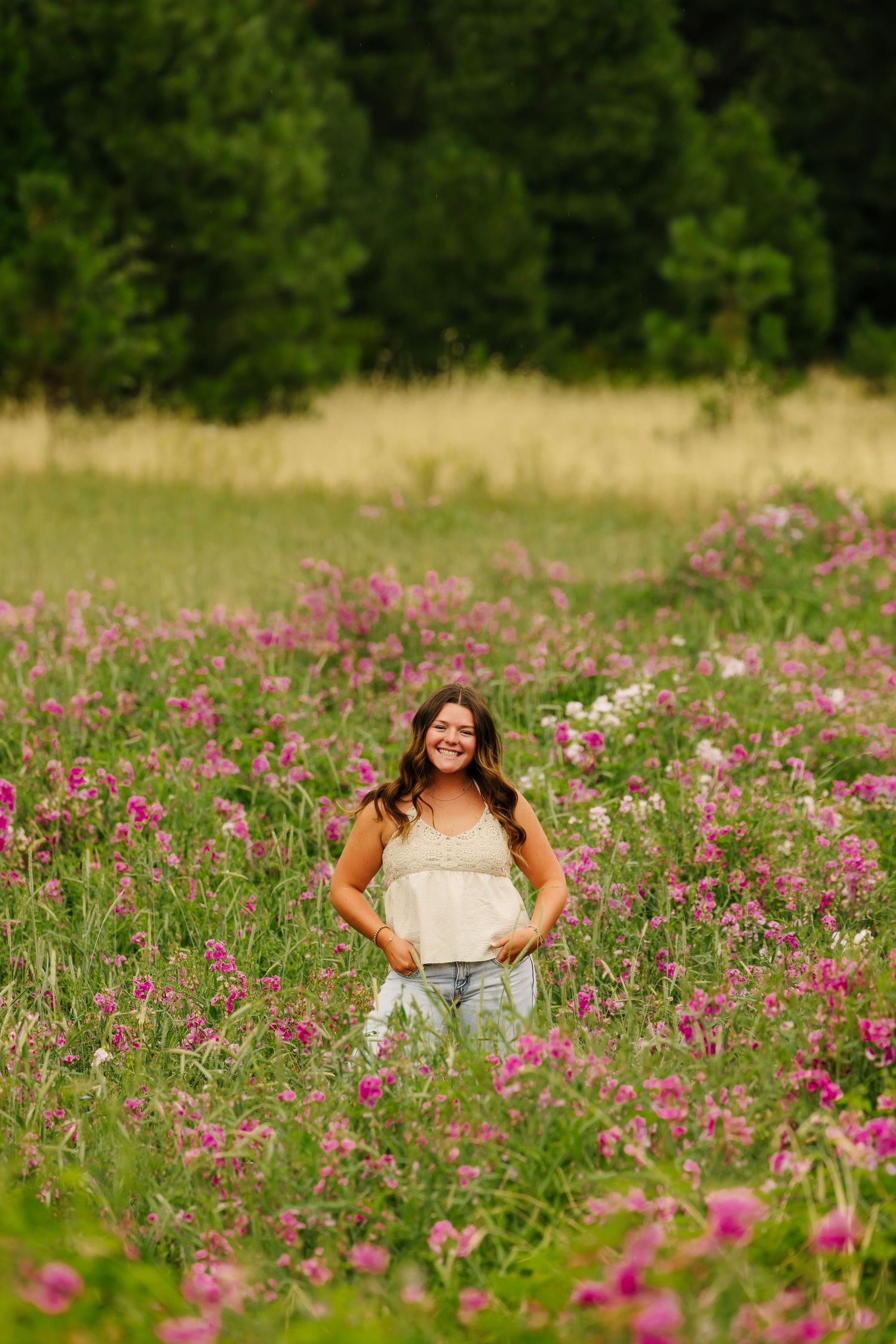 Young woman smiling in a vibrant wildflower meadow with pink blooms and lush green trees in the background.