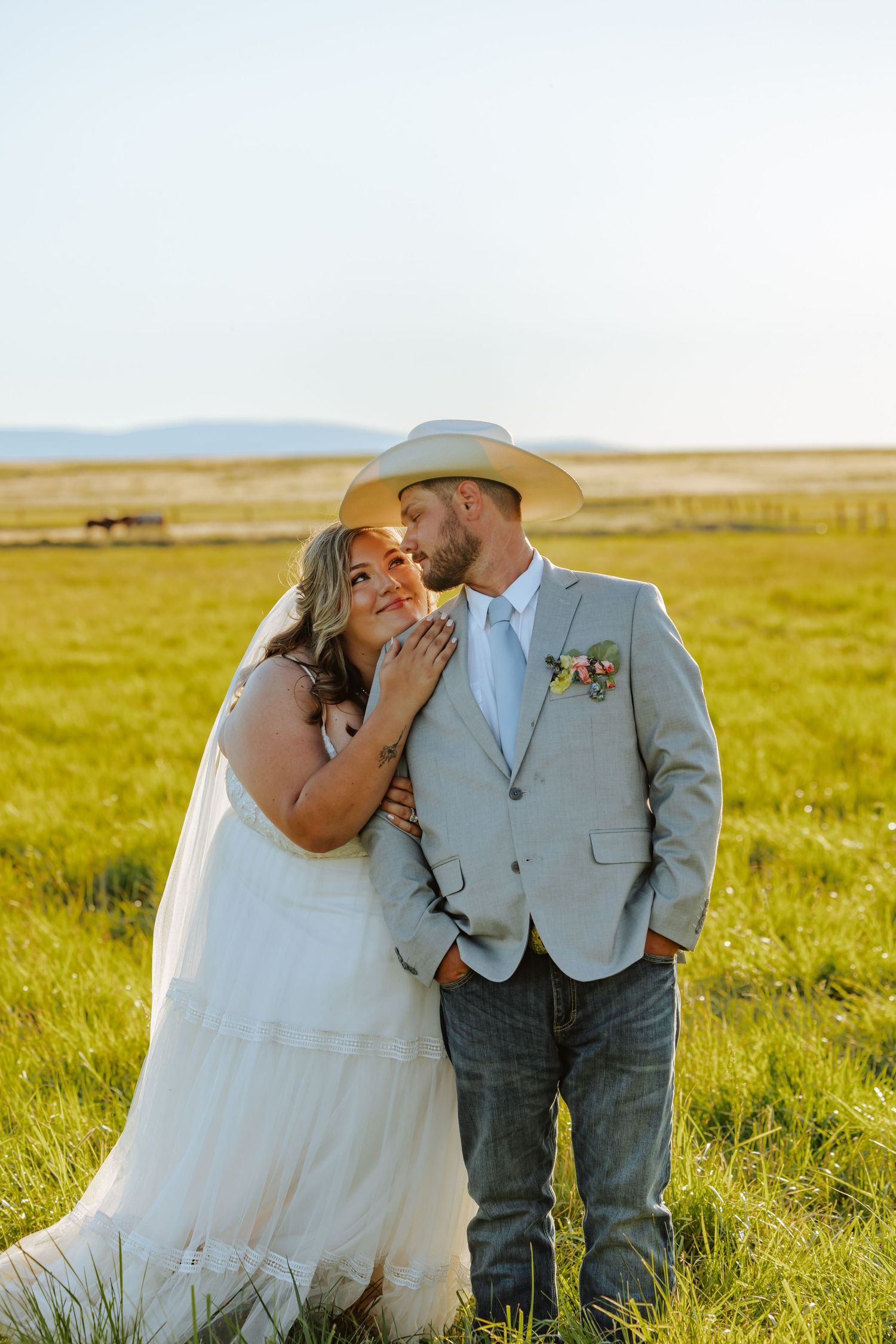 A bride and groom share a romantic moment in a sunlit green field, groom wearing a cowboy hat and boutonniere.