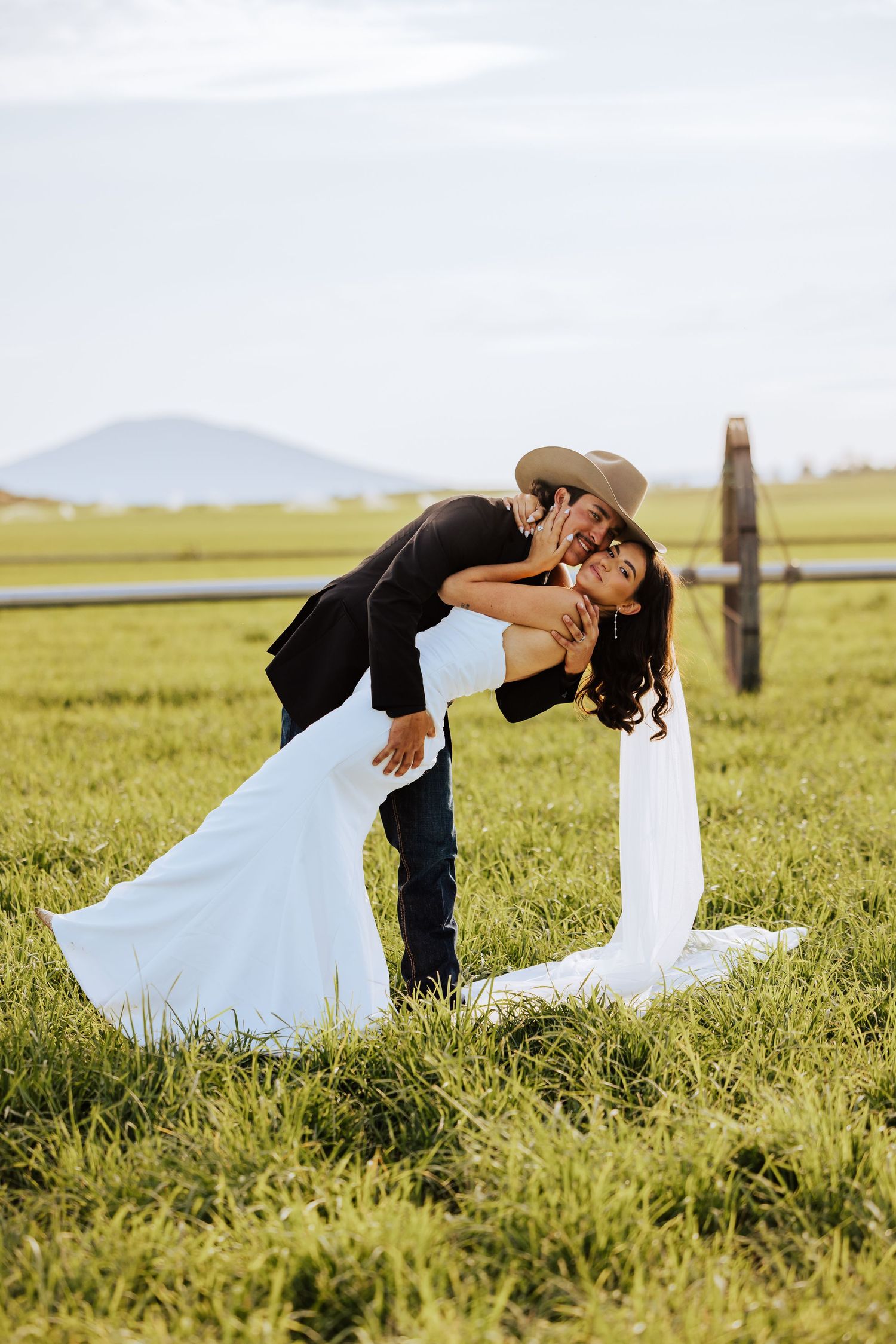 Groom in cowboy hat dips bride in white dress during romantic outdoor wedding photo on green field.