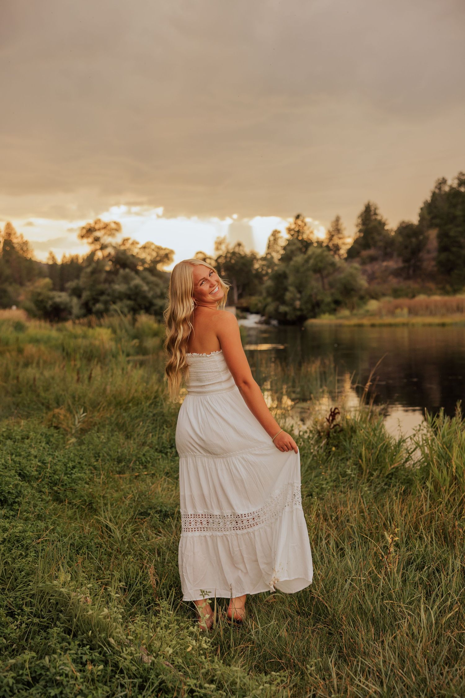 Person in white dress poses in tall grass by lake during sunset.