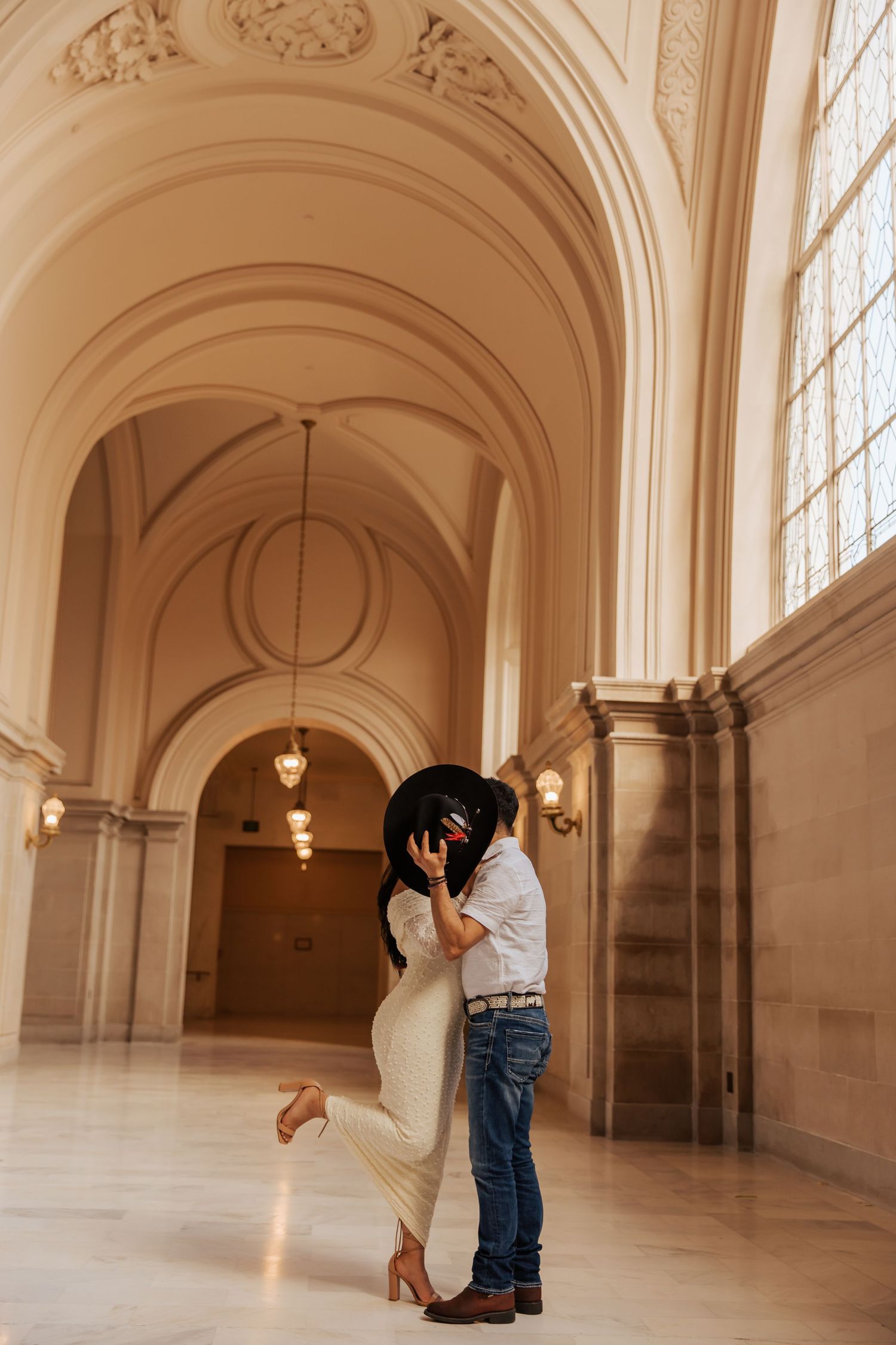 A romantic couple embraces in an elegant white marble hallway with arched ceilings and large windows.